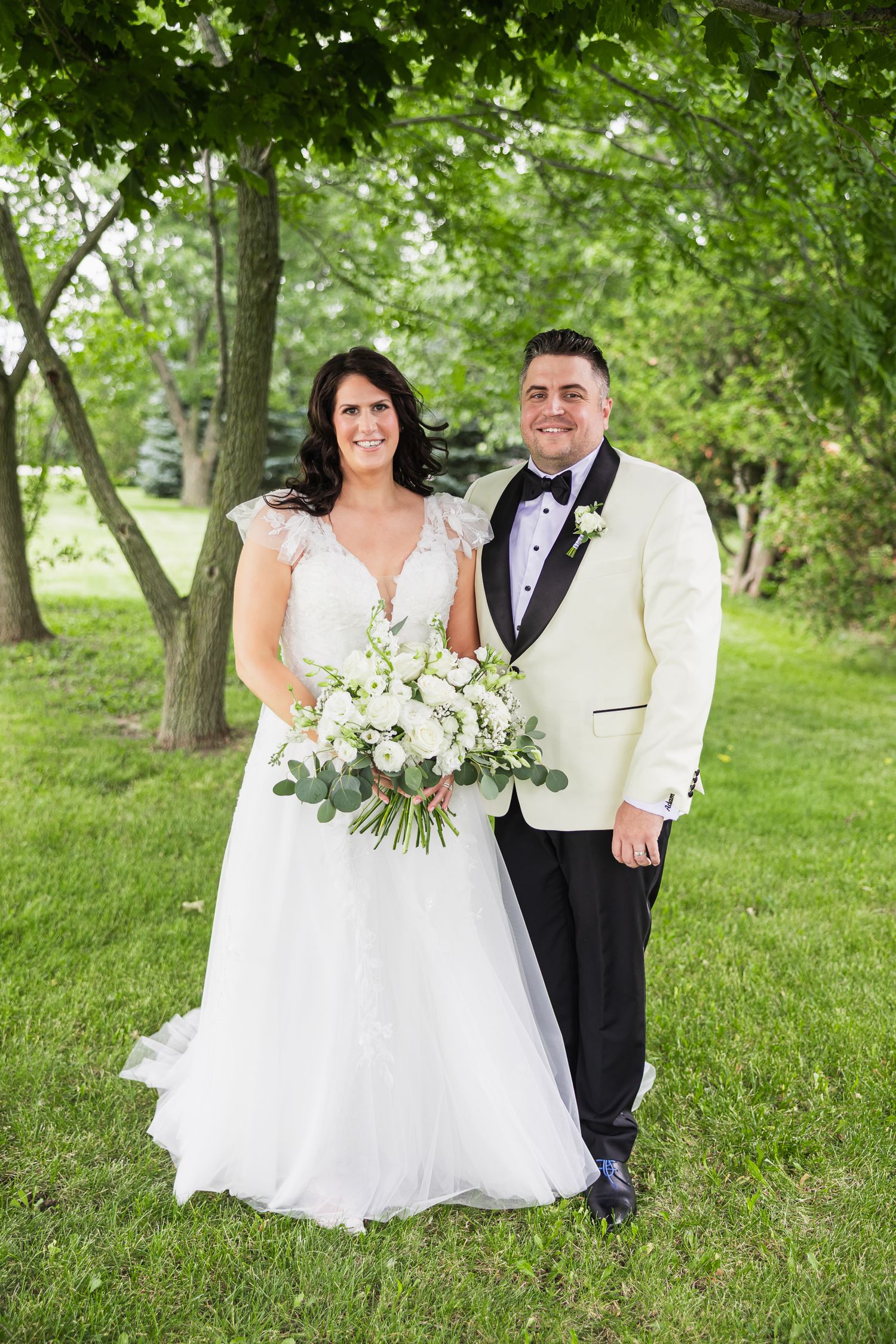 Bride in white lace gown holding bouquet stands with groom in cream tuxedo outdoors under green trees.