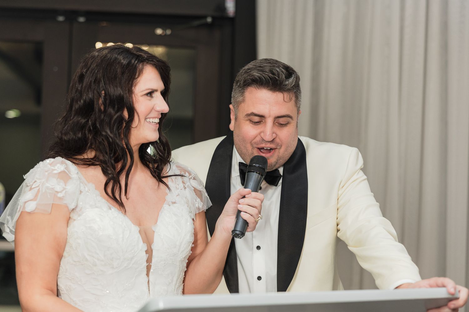 Bride in lace wedding gown and groom in white tuxedo singing together at wedding reception.