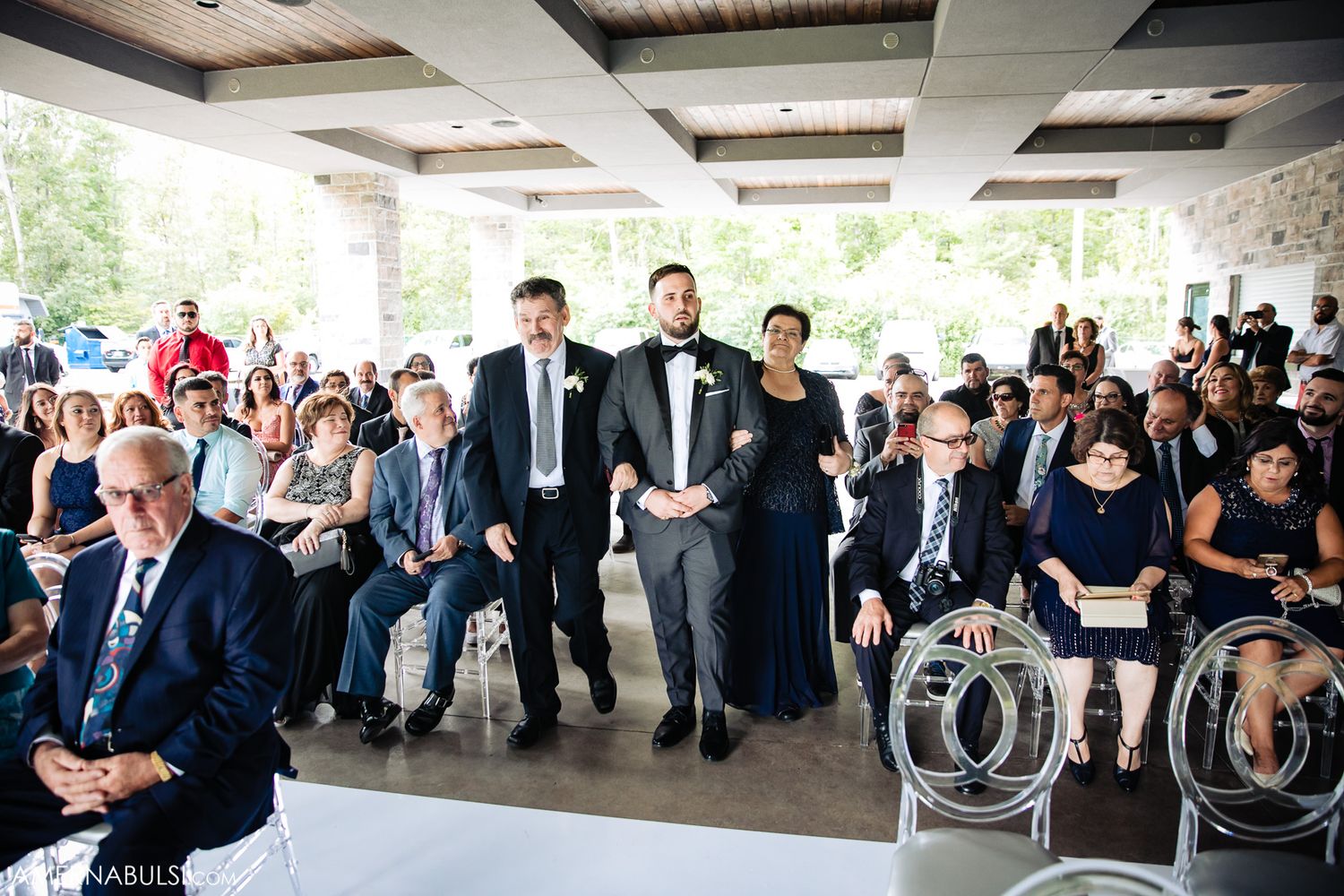 Wedding guests seated in rows at an indoor reception venue with rustic wooden beam details.