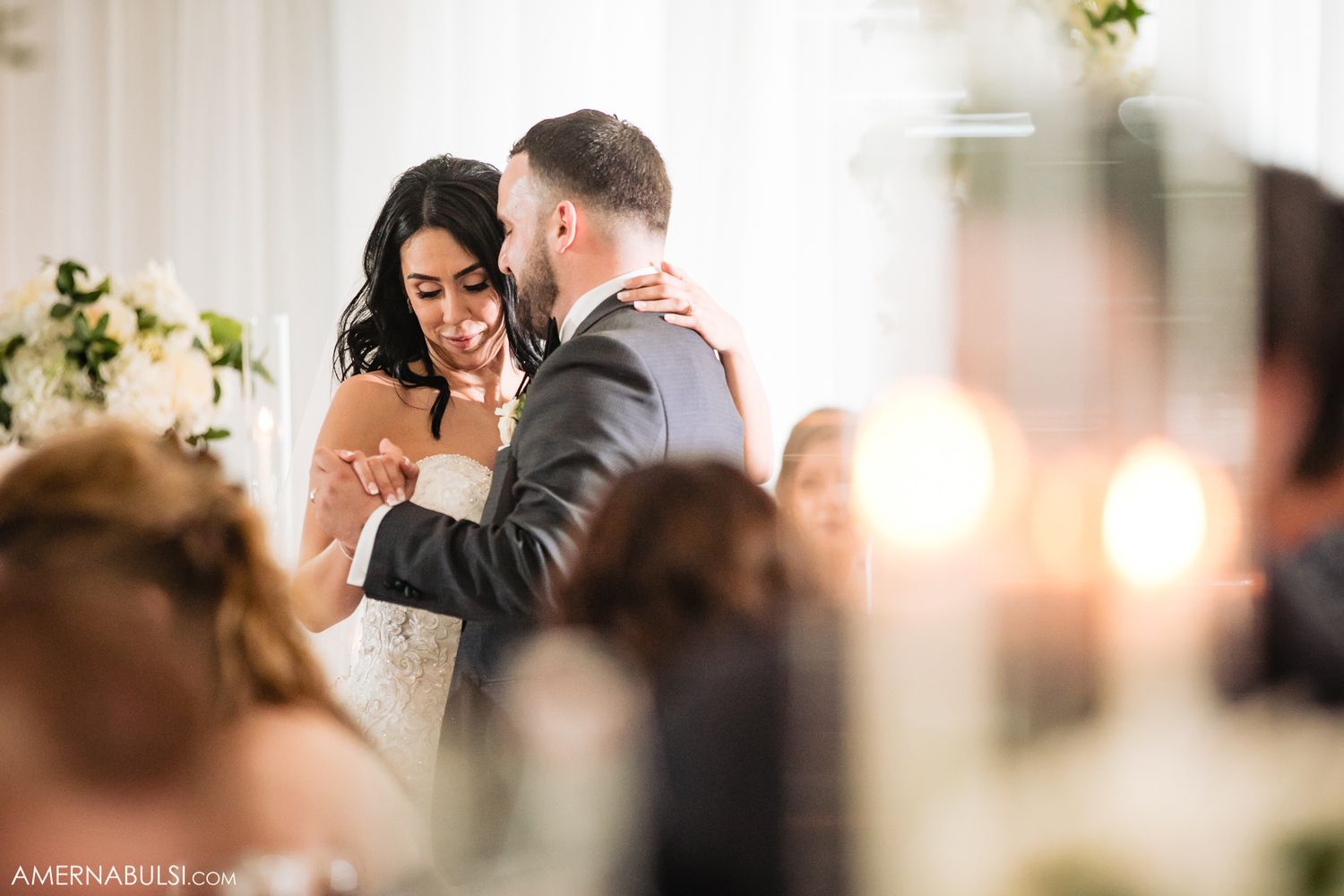 A couple shares a tender moment during their wedding reception as guests look on in soft lighting.
