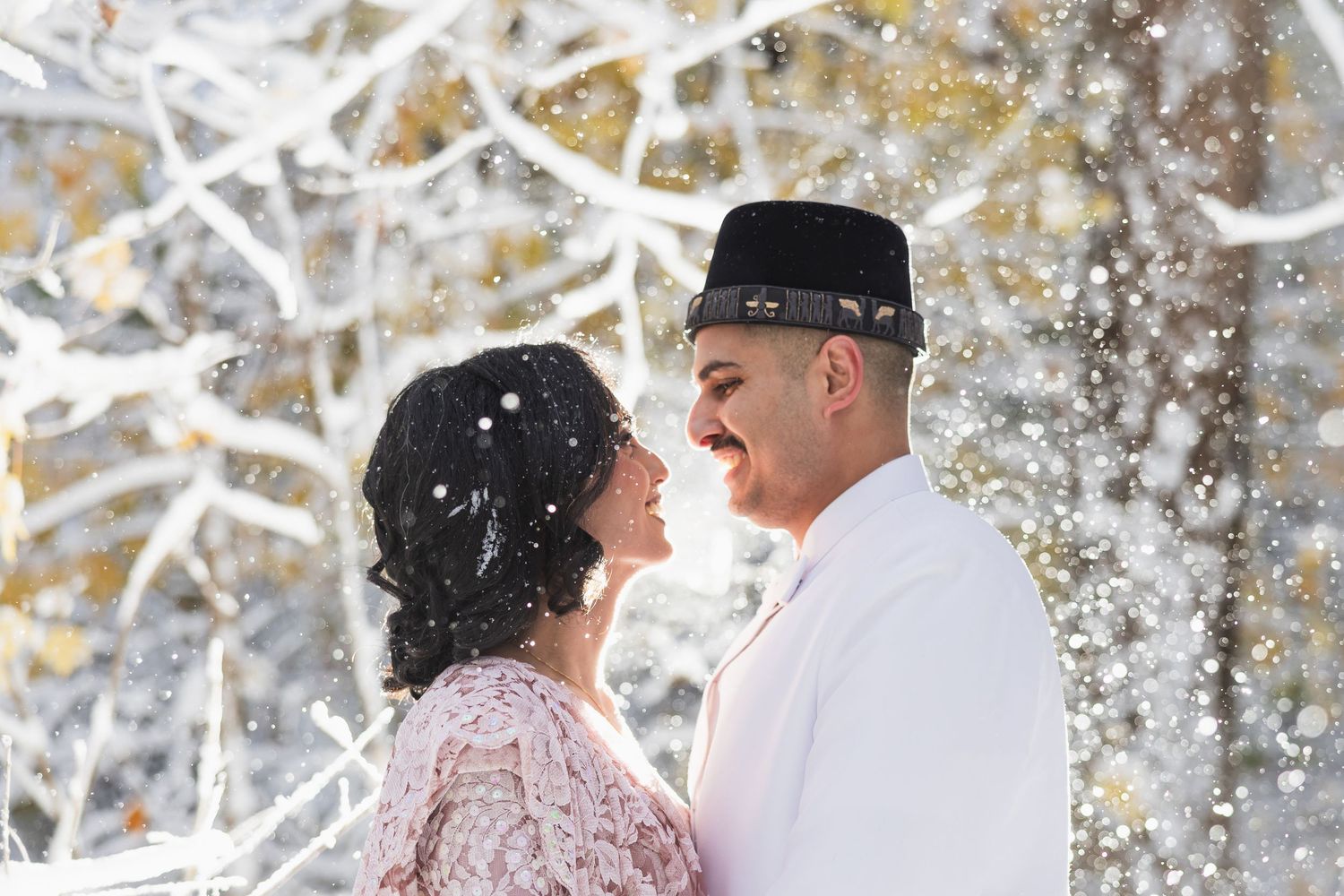 A couple shares a romantic moment in winter as snow falls around them against a snowy white backdrop.