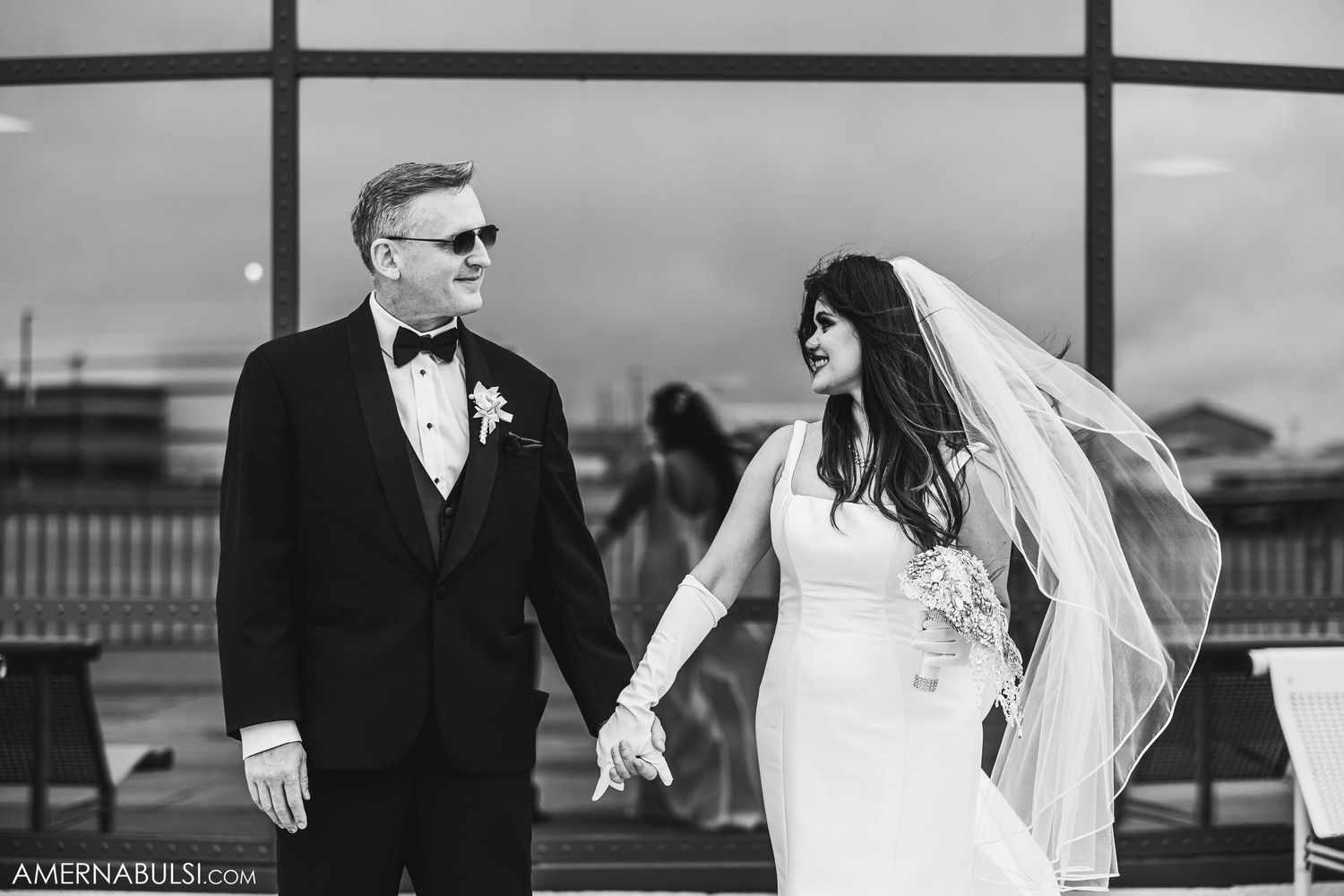 A wedding couple shares an intimate moment in formal attire against a modern window backdrop.