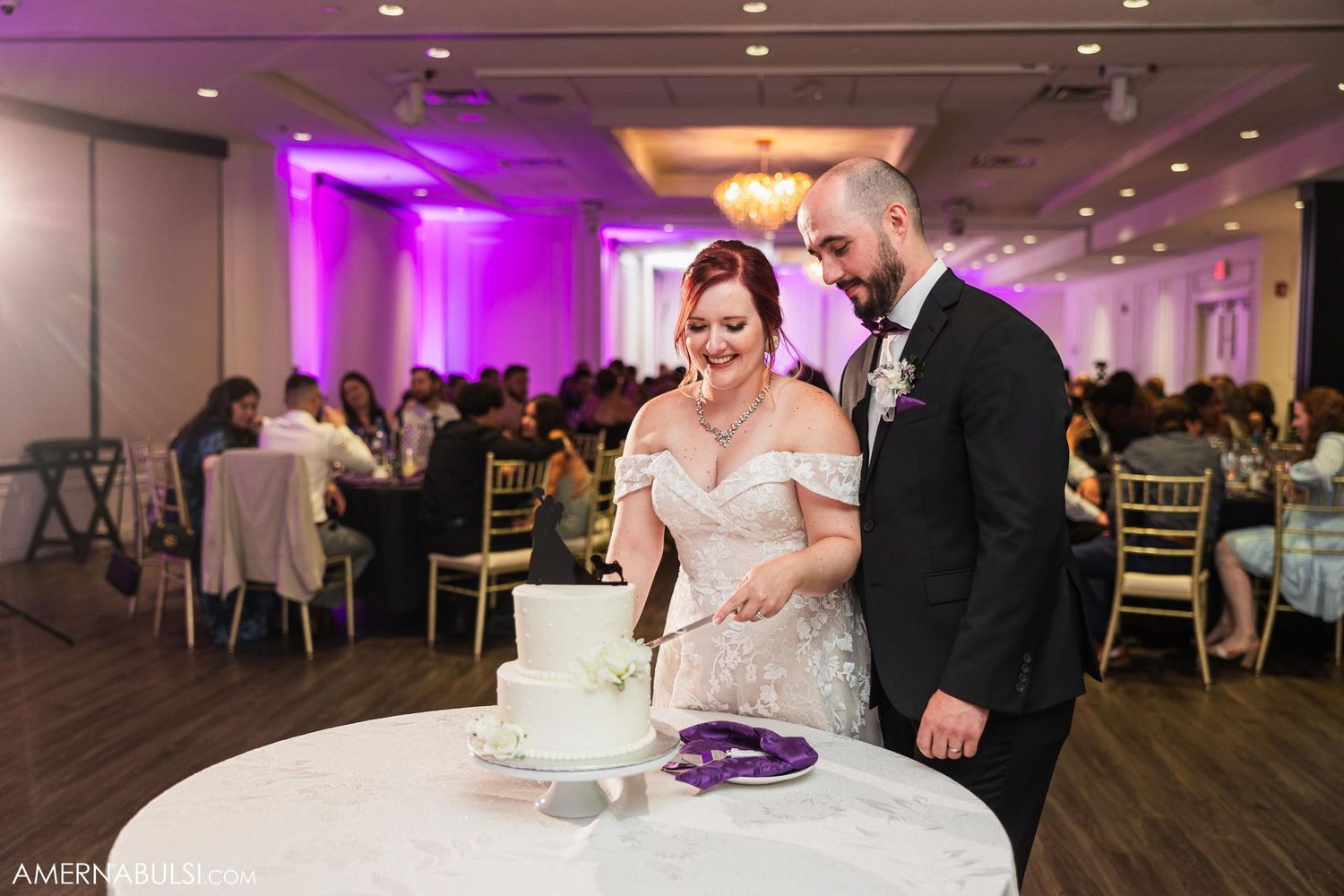 Newlyweds standing at cake table during reception with purple uplighting.