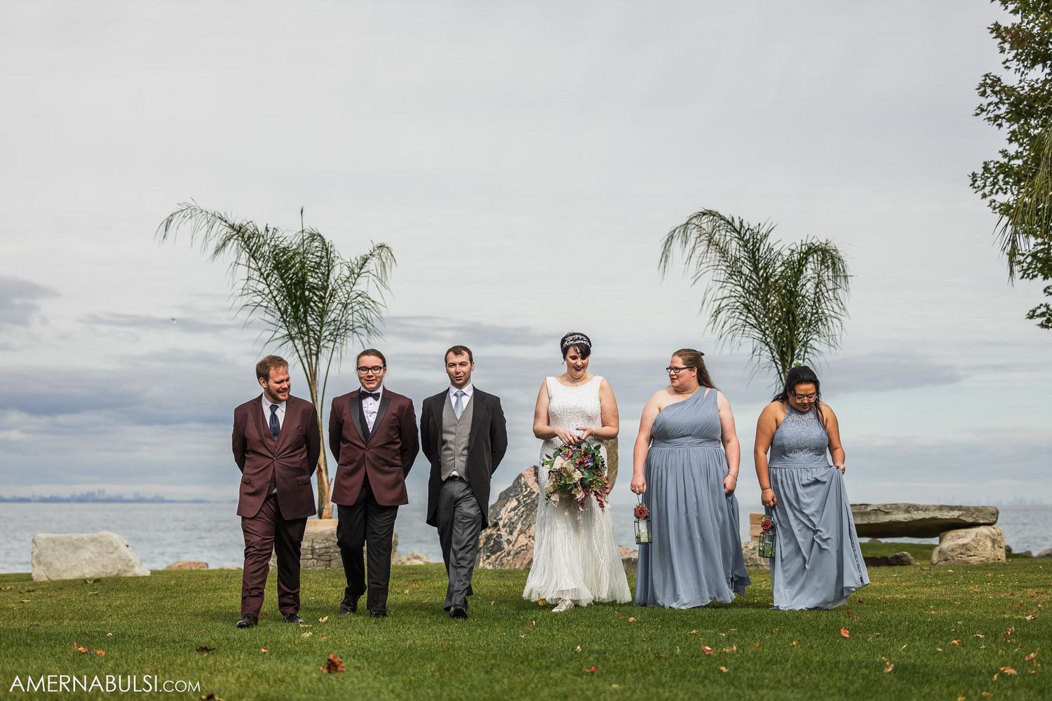 Wedding party lined up outdoors with palm trees and ocean view in background.