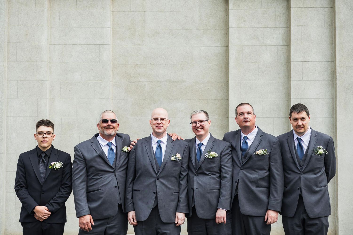 Wedding party members in dark suits stand in a row against white stone wall.