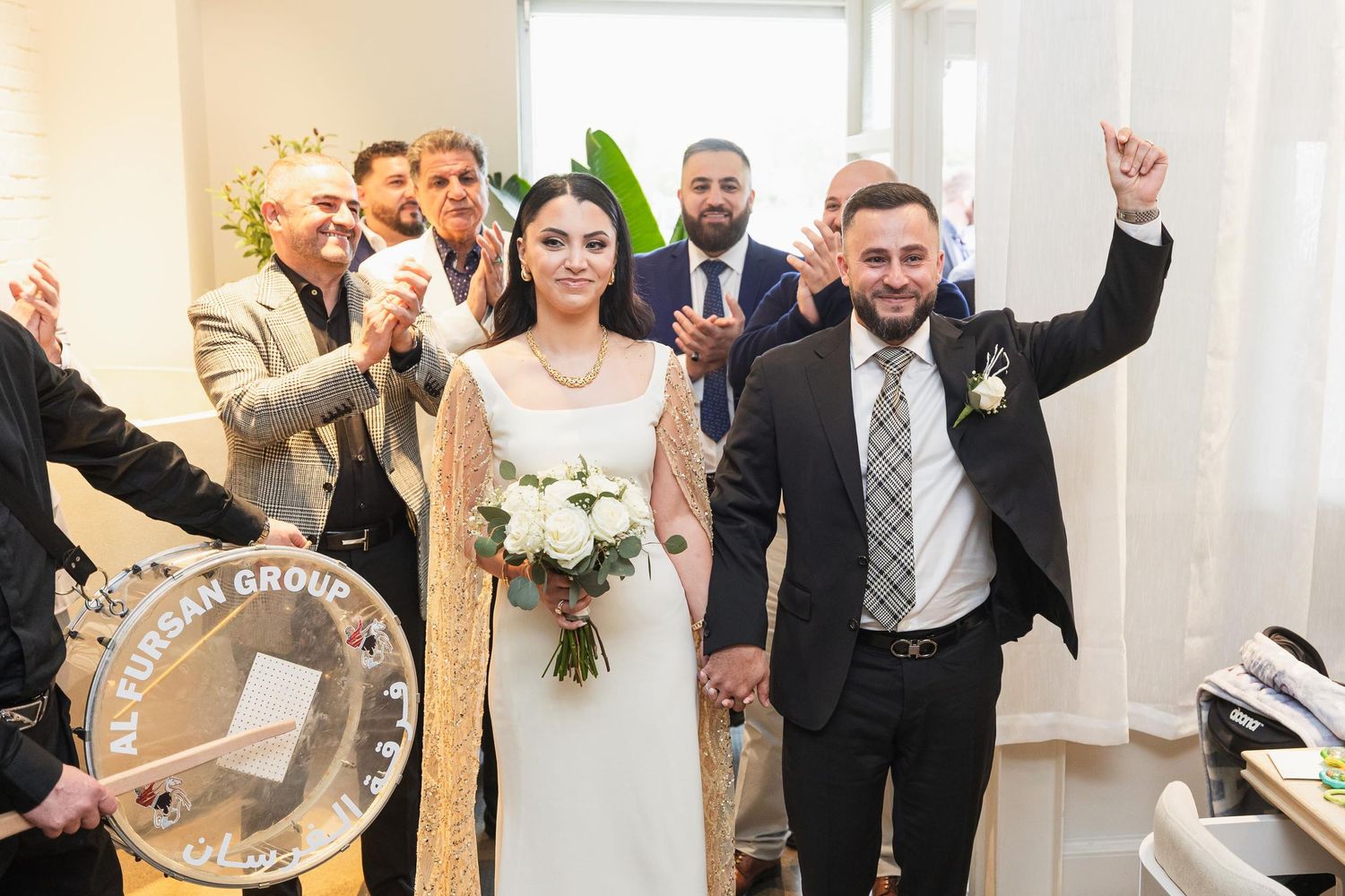 Wedding party celebrates joyfully after ceremony in bright indoor venue with raised arms and white decor.