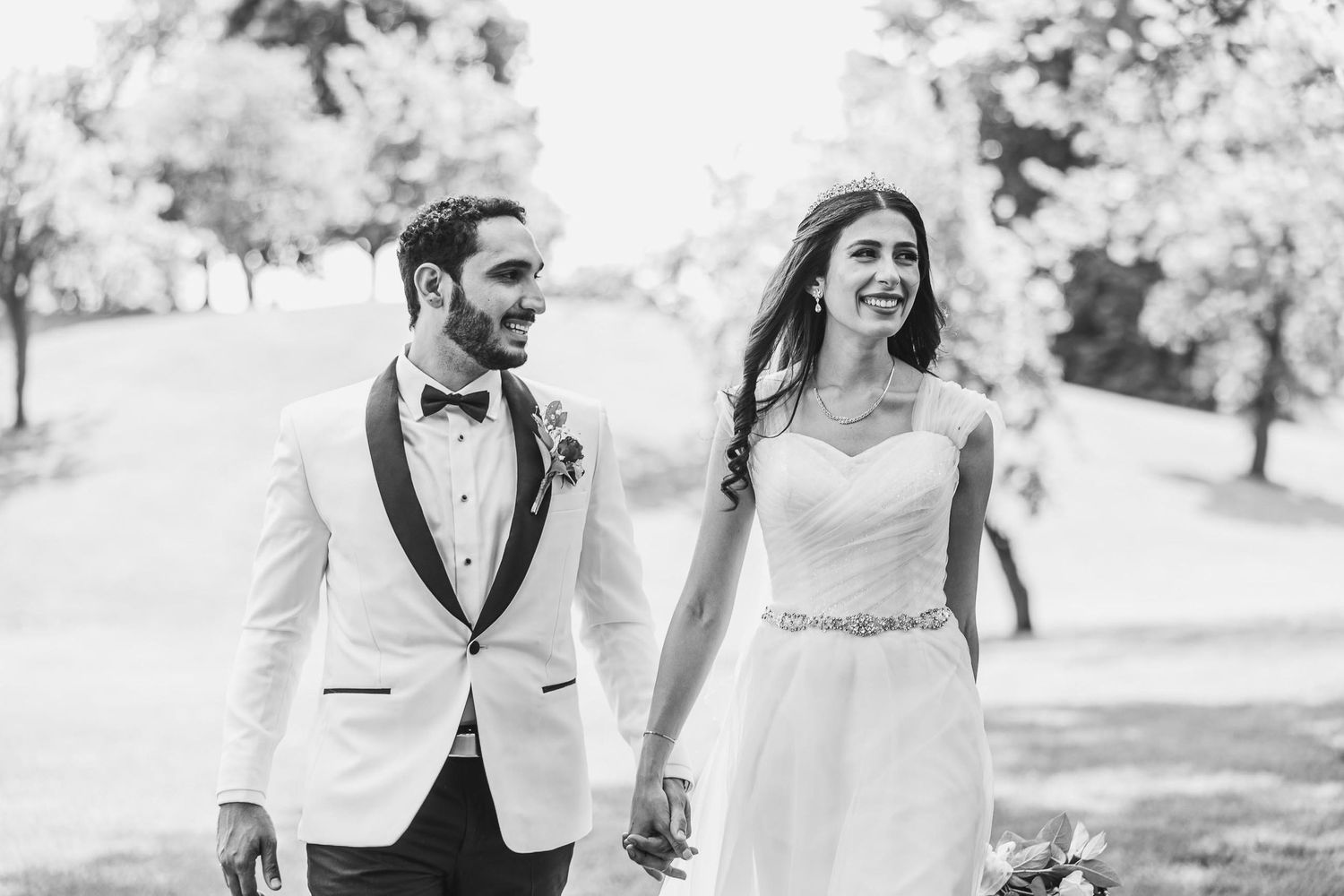 Happy couple walking hand in hand through sunny park in wedding attire.