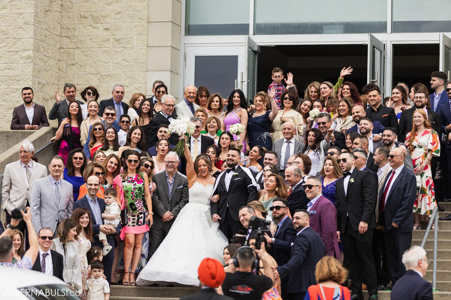 Large wedding group photo with guests gathered on steps outside modern building.