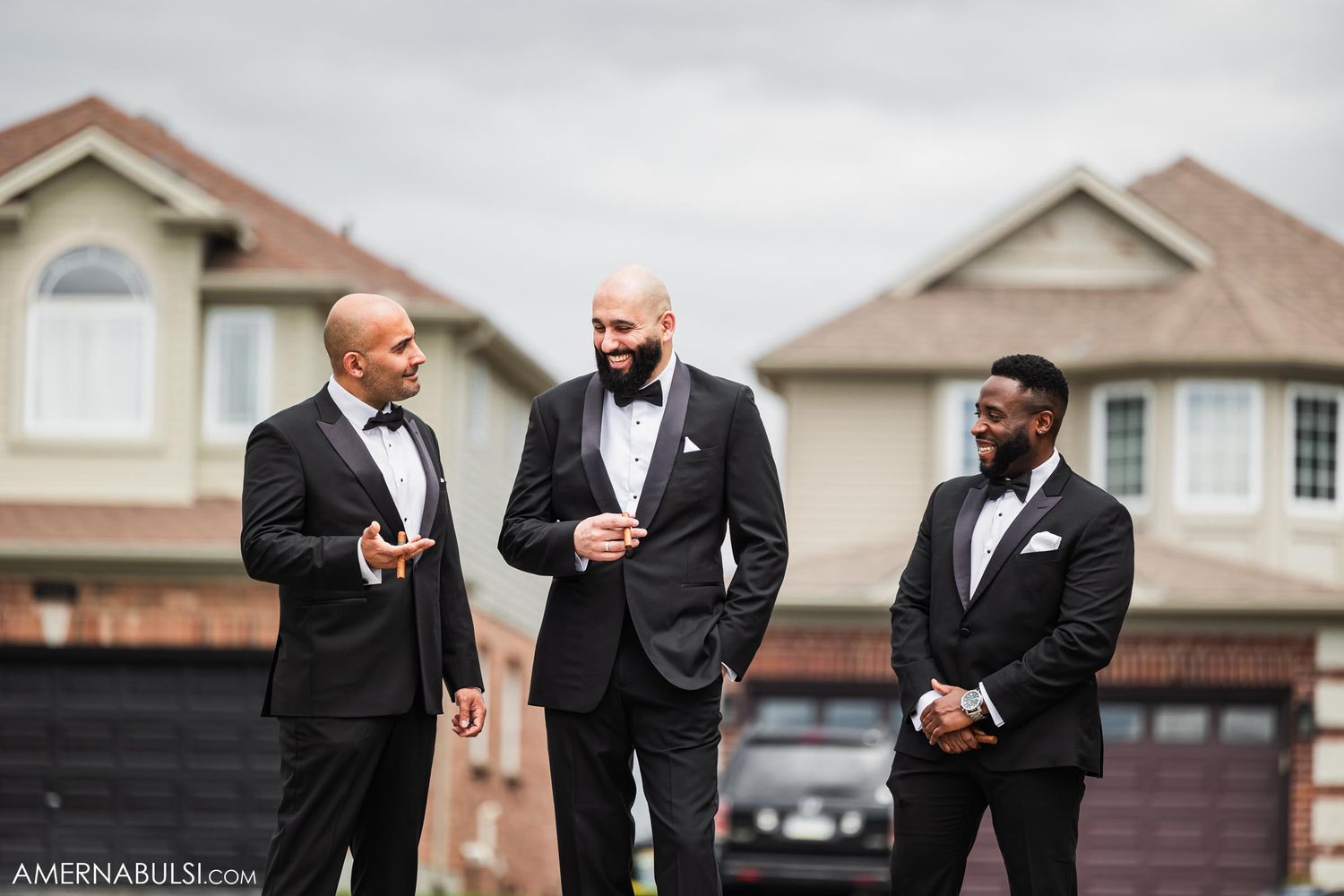 Three people in formal tuxedos standing and conversing outside suburban homes.