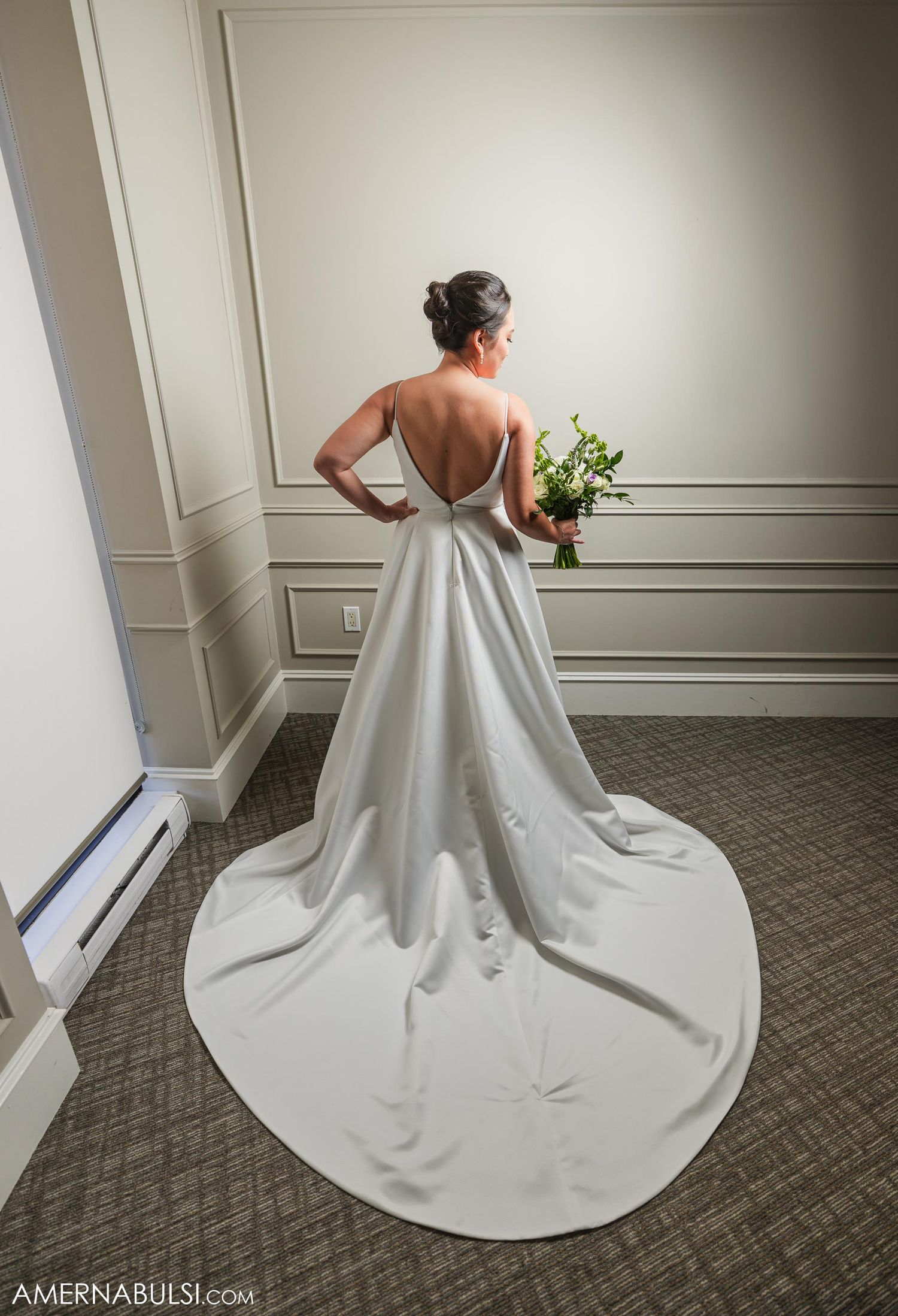 Back view of bride in classic white wedding gown with dramatic train against neutral wall.