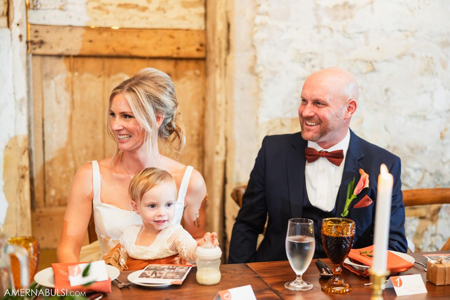 Newlyweds and young child enjoy dinner at rustic wedding reception head table.