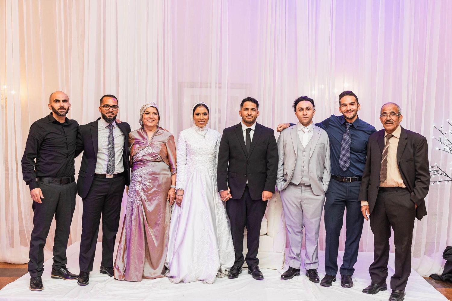 Group photo of wedding party members in formal attire standing against white backdrop with soft lighting.