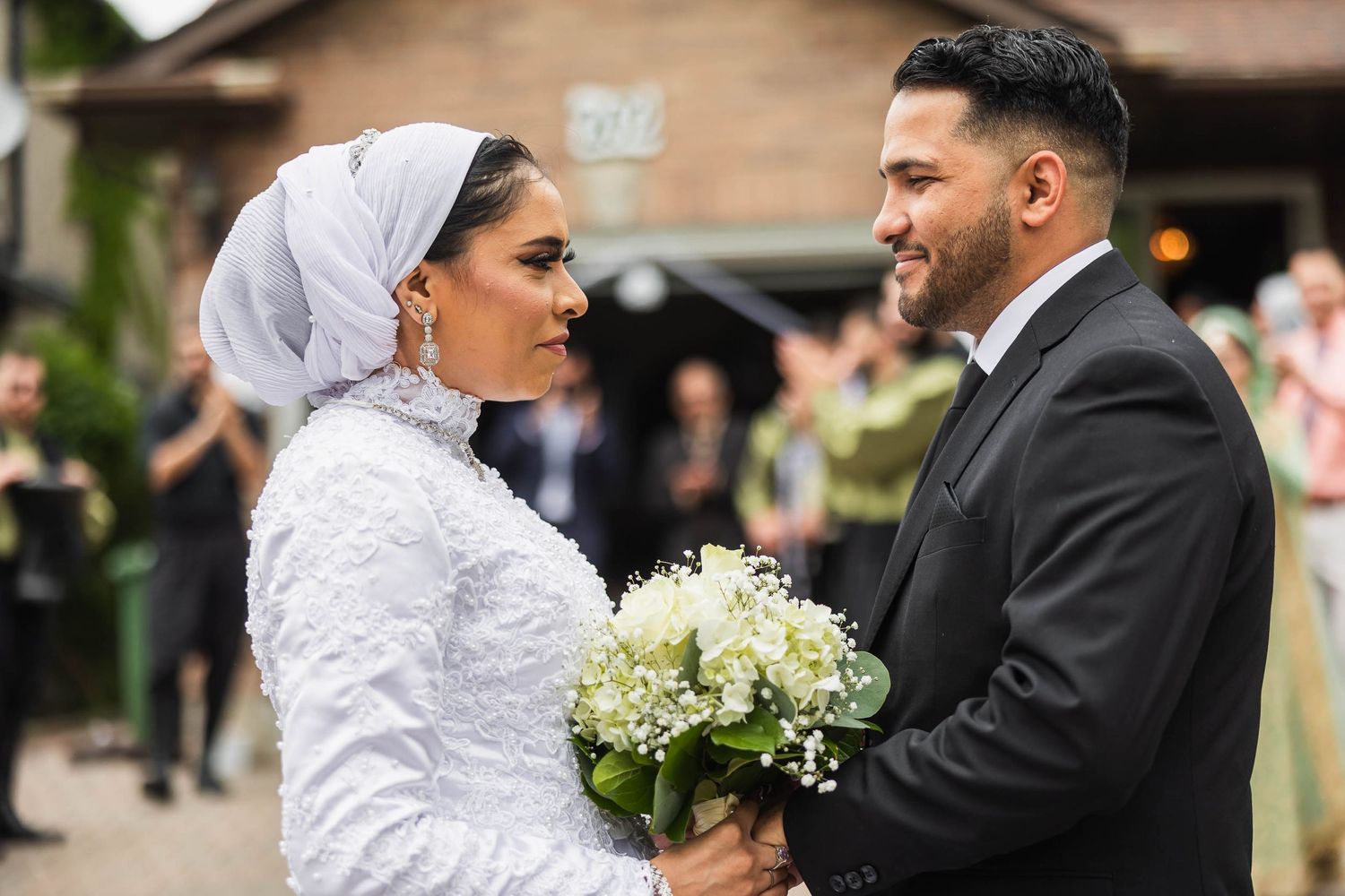 A bride in a white lace hijab dress holds a bouquet while standing with her groom at their wedding.