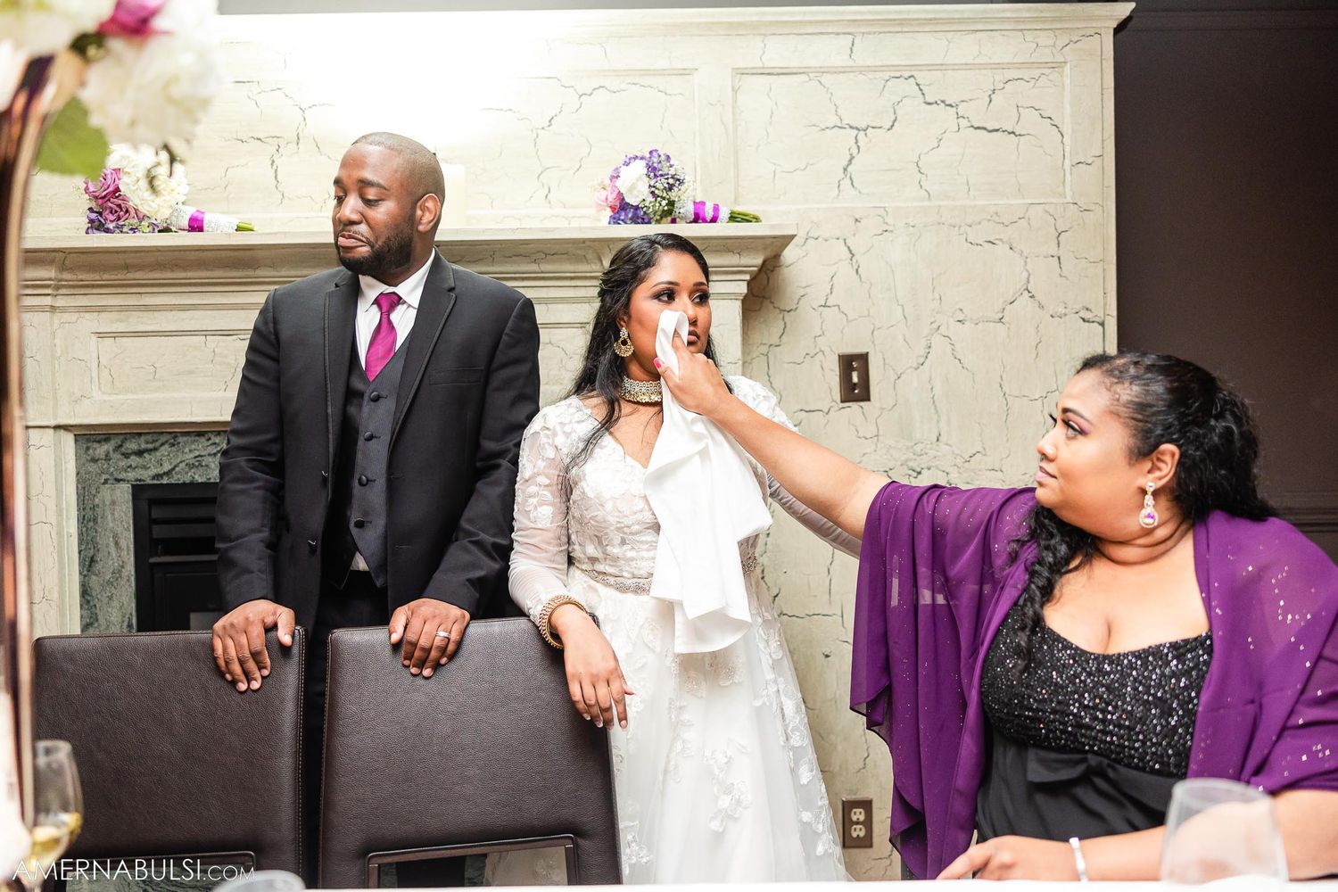 Wedding celebration scene with guests sharing laughter by decorative fireplace.