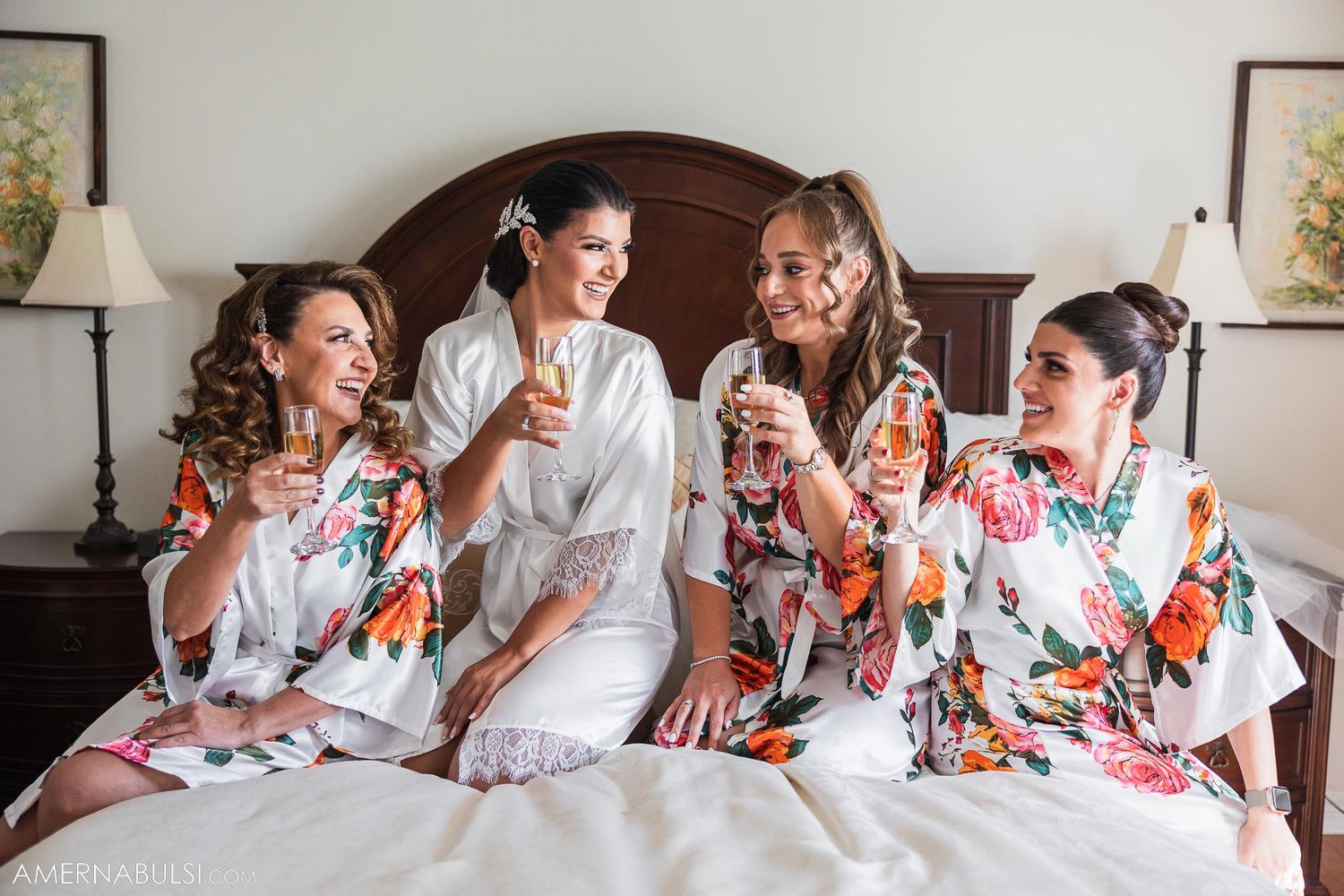 A group in matching floral robes share champagne and laughter while sitting on a bed.