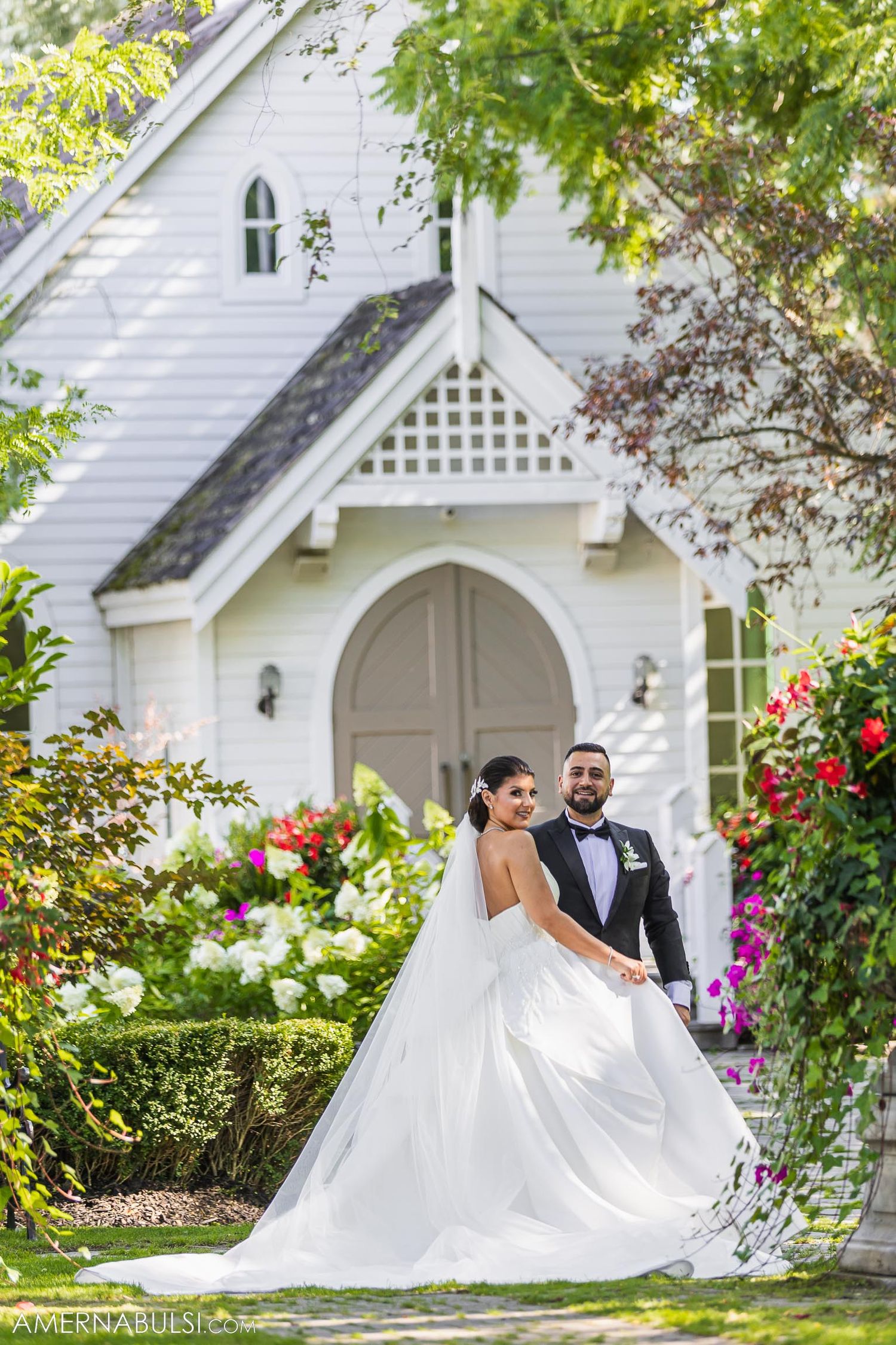 A couple poses in front of a white chapel with lush garden landscaping.