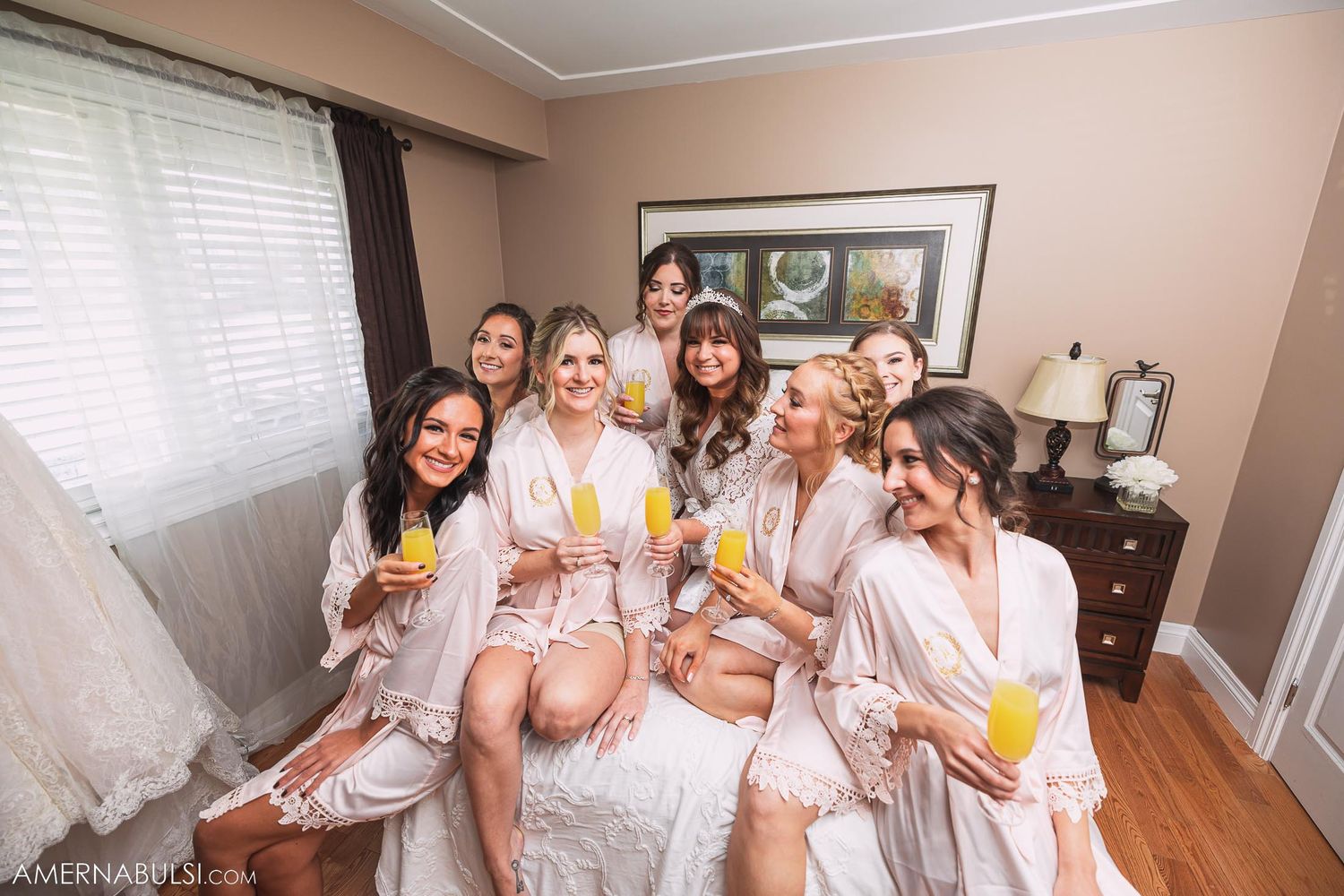 Bridesmaids in matching robes share morning mimosas while sitting on a bed during wedding preparations.