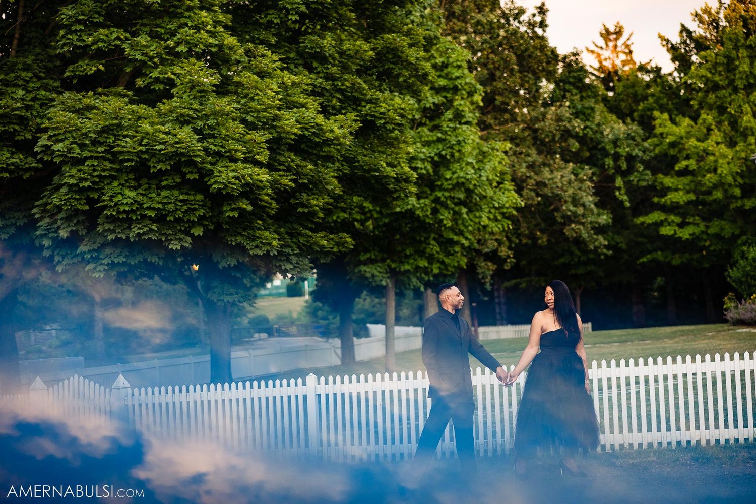 Couple in formal wear pose by white picket fence with atmospheric smoke effect.