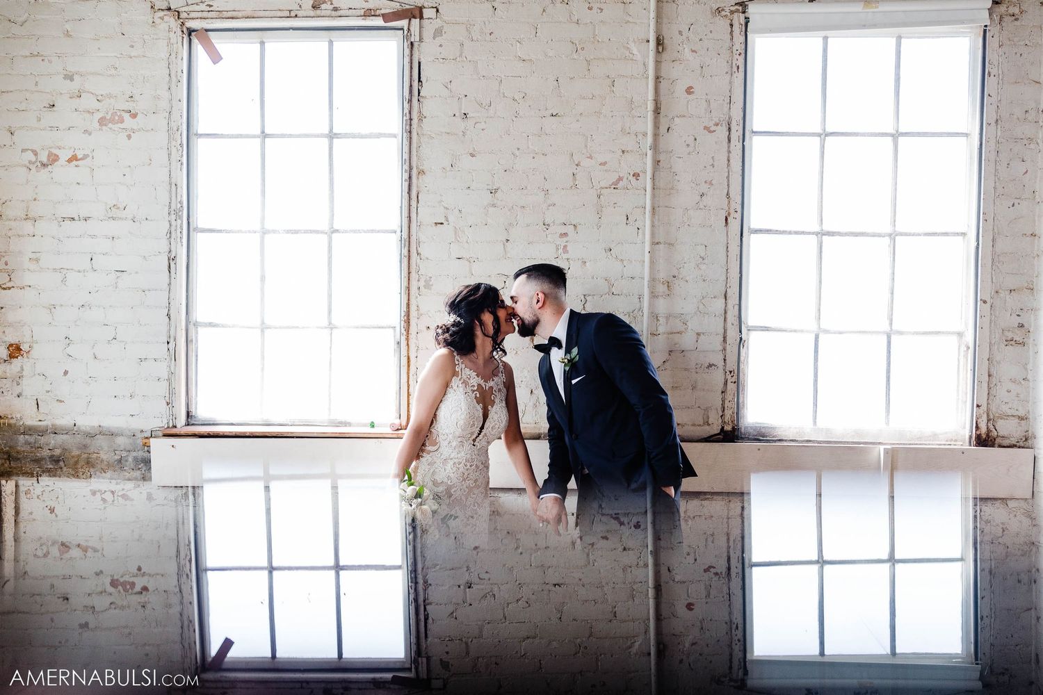 Artistic reflection shot of couple in formal wear against white brick wall and windows.