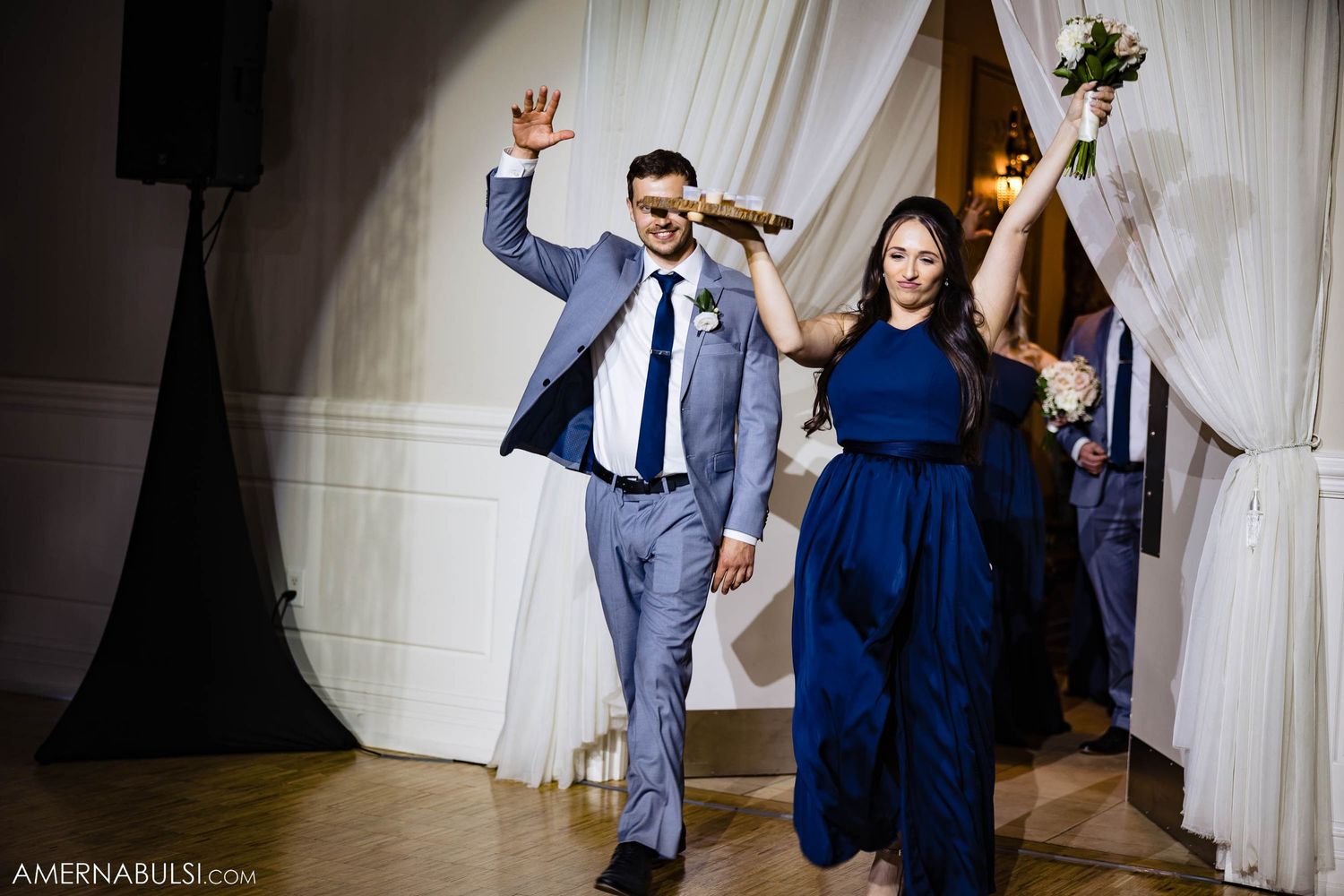 Wedding attendants make their grand entrance at reception, dancing enthusiastically in navy and grey formal wear.