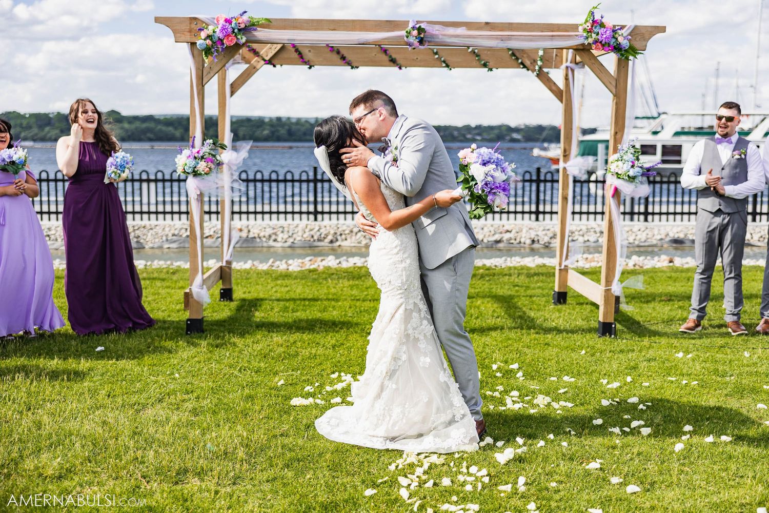 Newlyweds share romantic moment under rustic wooden arch decorated with purple flowers by waterfront.