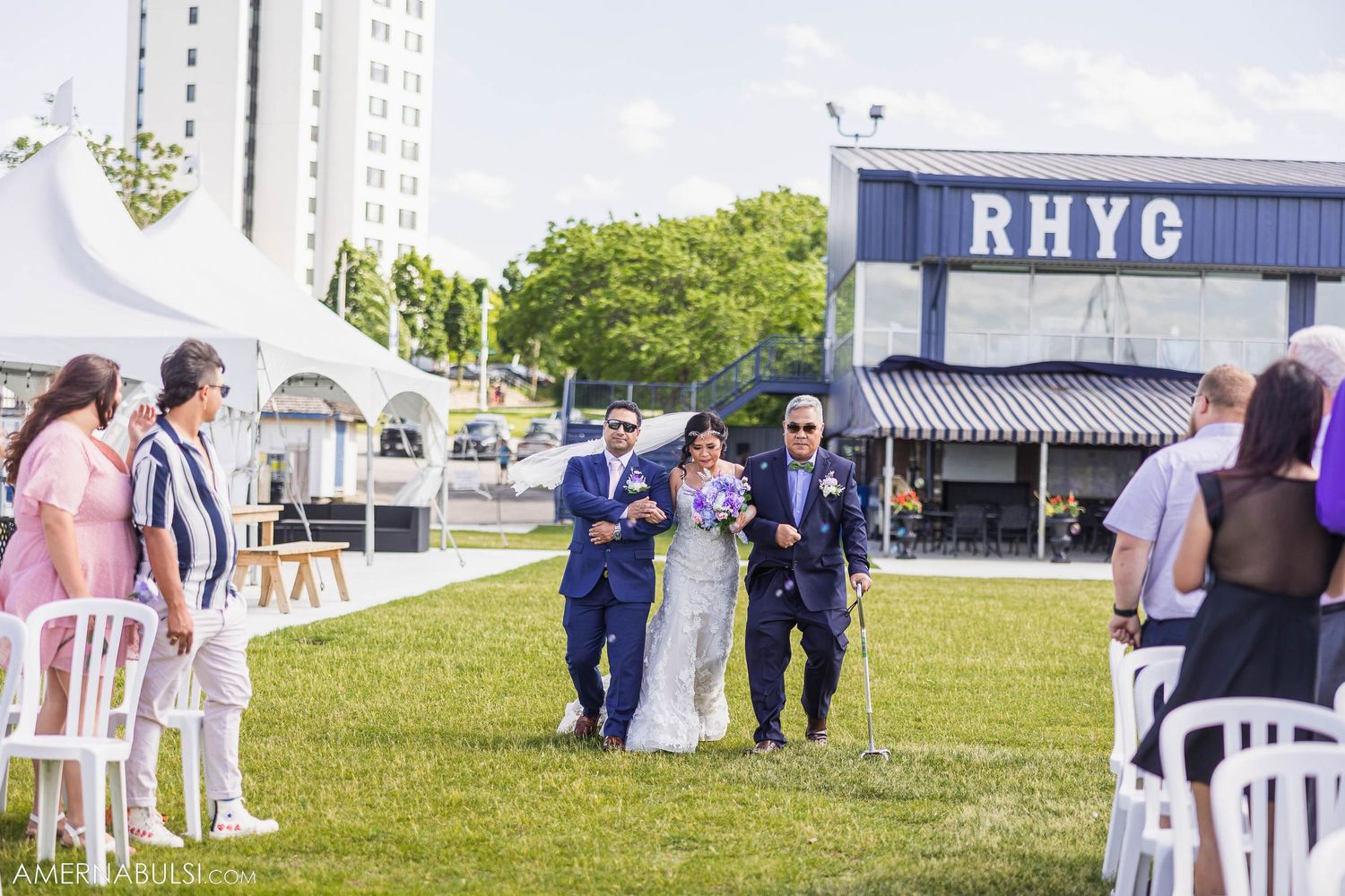 Wedding guests walking between white chairs on grassy venue with RHYC building in background.