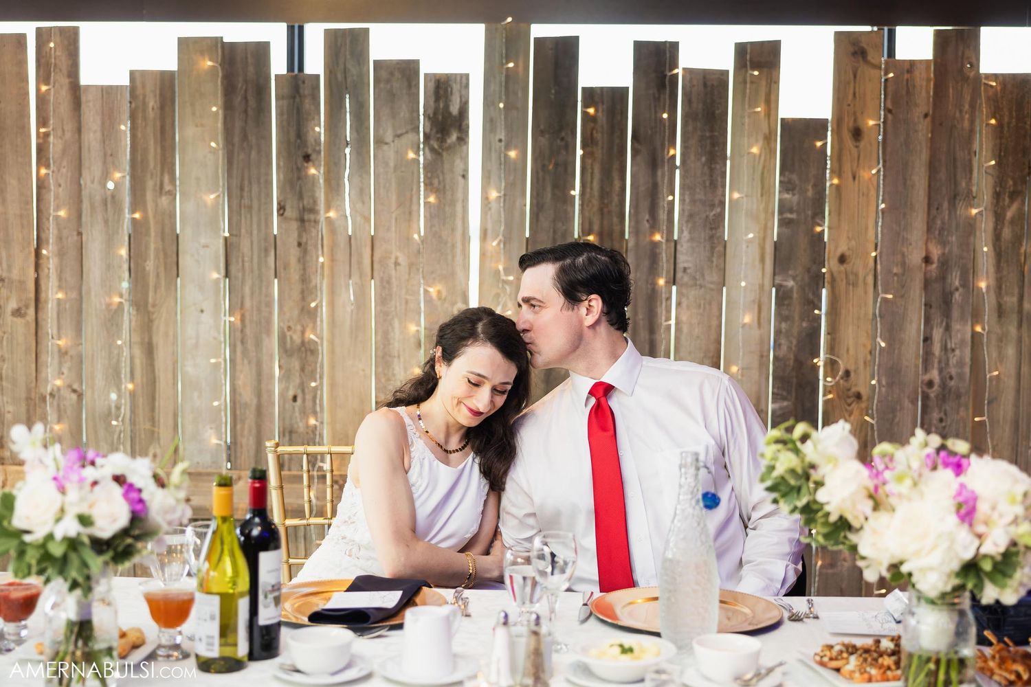 Intimate moment at rustic wooden reception table with flowers and wine bottles.