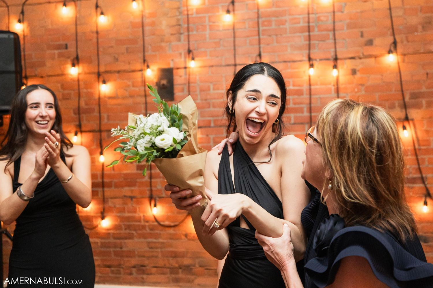 Joyful celebration moment with bouquet of flowers against exposed brick wall with string lights.