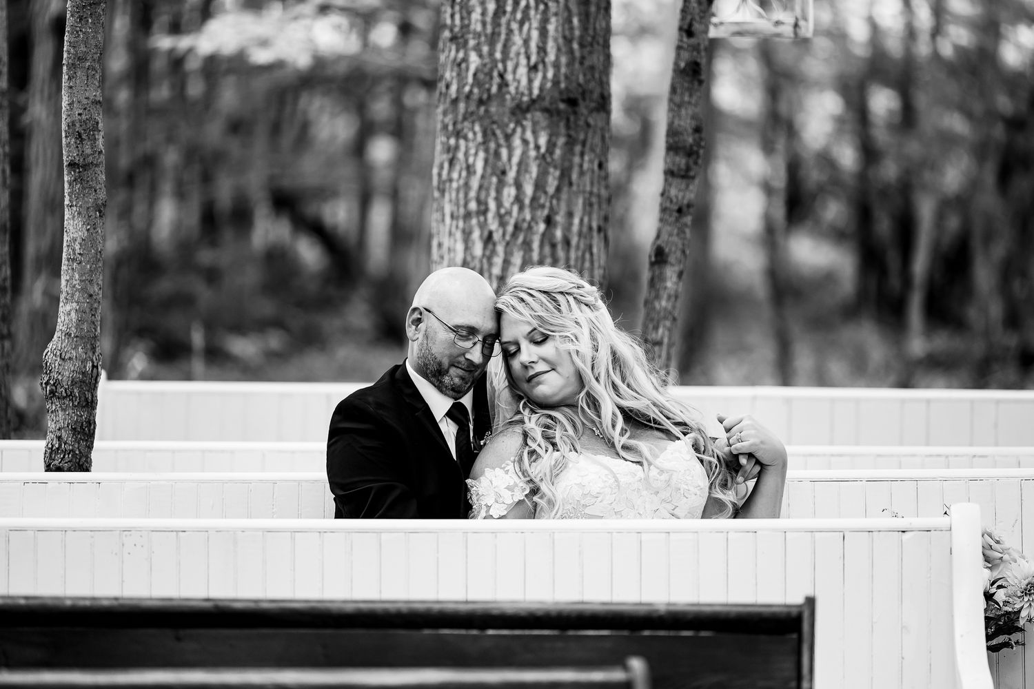 Black and white photo of wedding party standing along a white bench in a wooded outdoor setting.