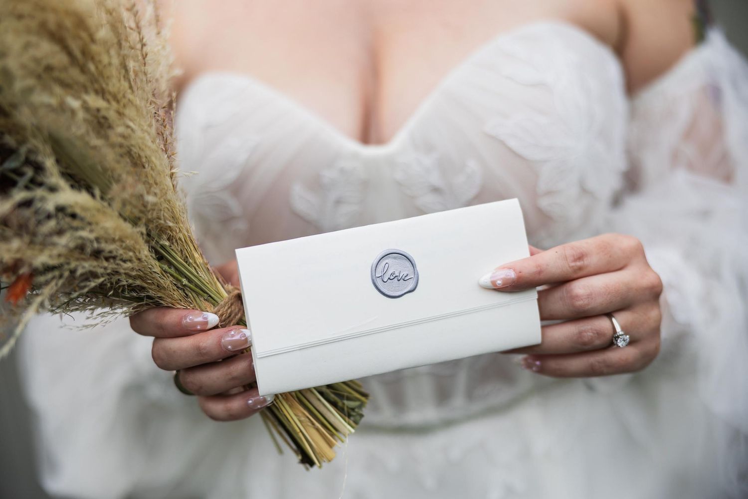 Close up of bride holding white envelope with wax seal and dried flowers.