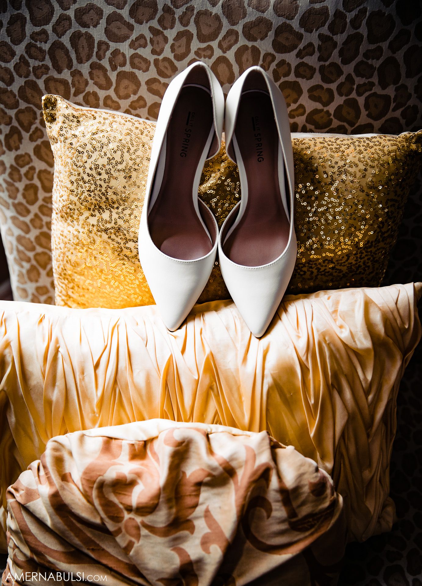 Wedding shoes displayed on silky fabric against a patterned backdrop with golden lighting.