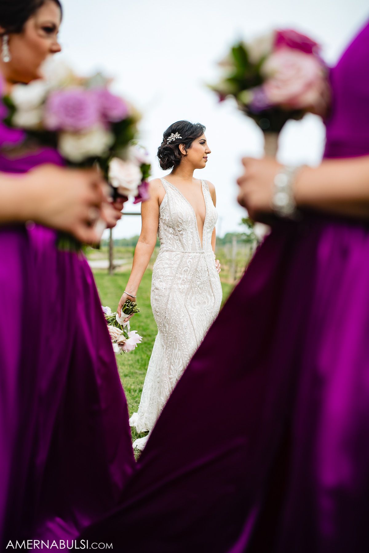 Elegant bride in white lace dress stands among bridesmaids wearing deep purple gowns.