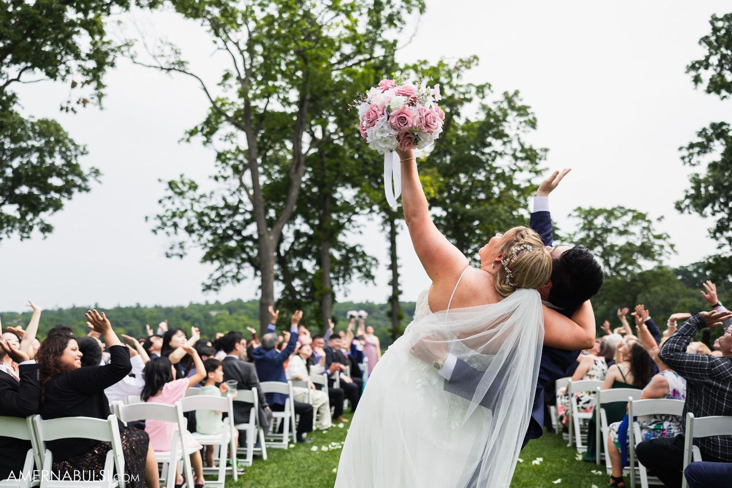 Newlyweds celebrate with guests during recessional as bride raises bouquet triumphantly outdoors.