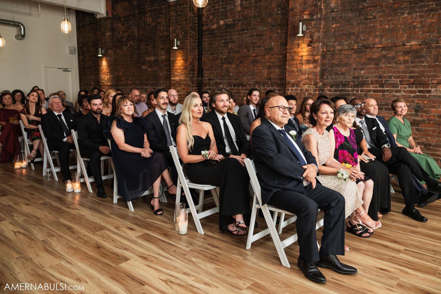 Wedding guests seated in white folding chairs watching ceremony in rustic brick-walled venue.