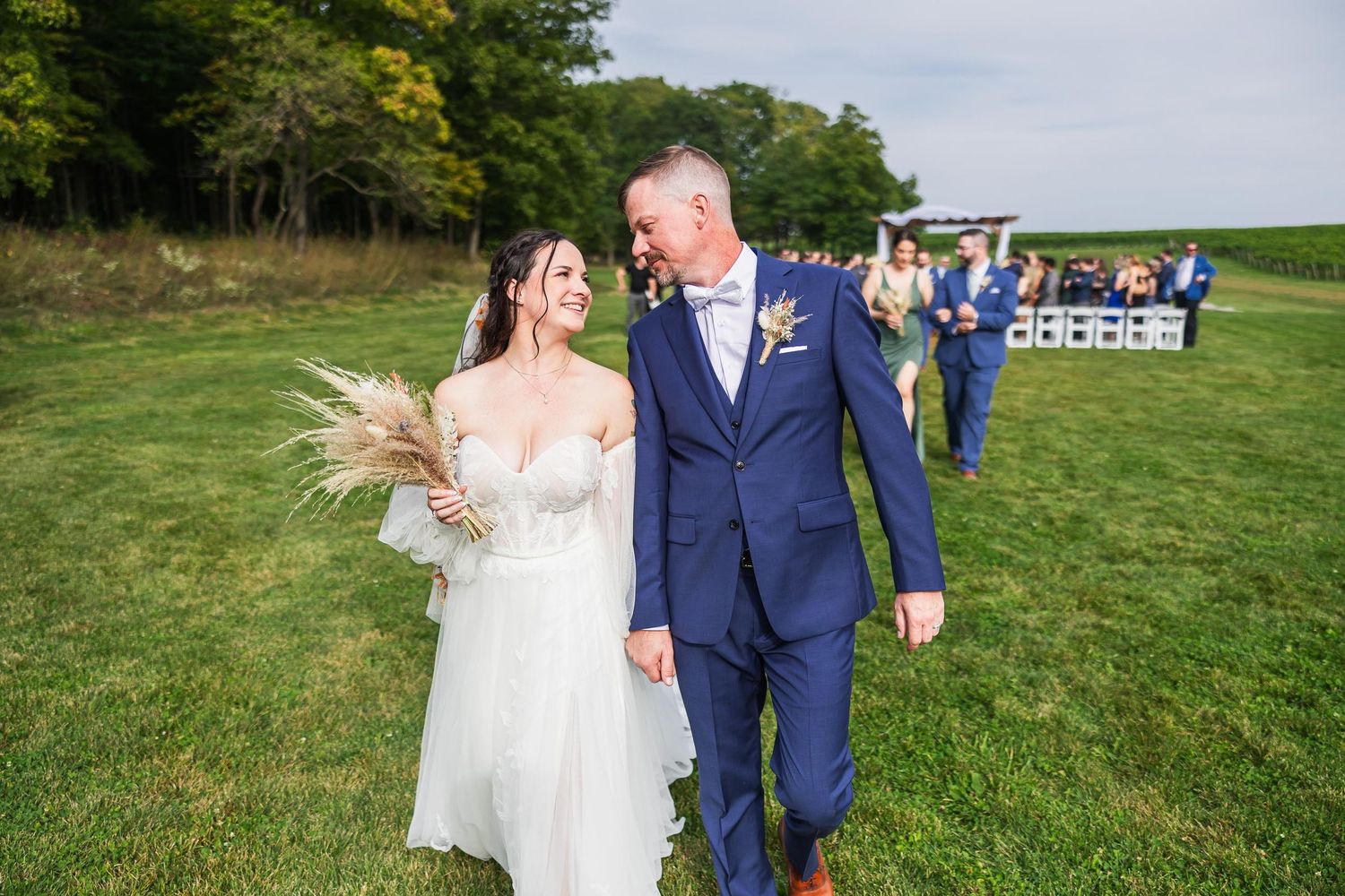 Newlyweds walking together on green lawn during outdoor countryside wedding celebration.