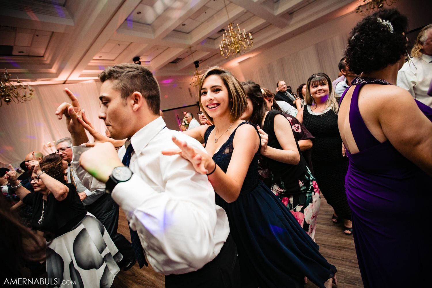 Wedding guests dancing energetically on a crowded ballroom dance floor under elegant chandeliers.