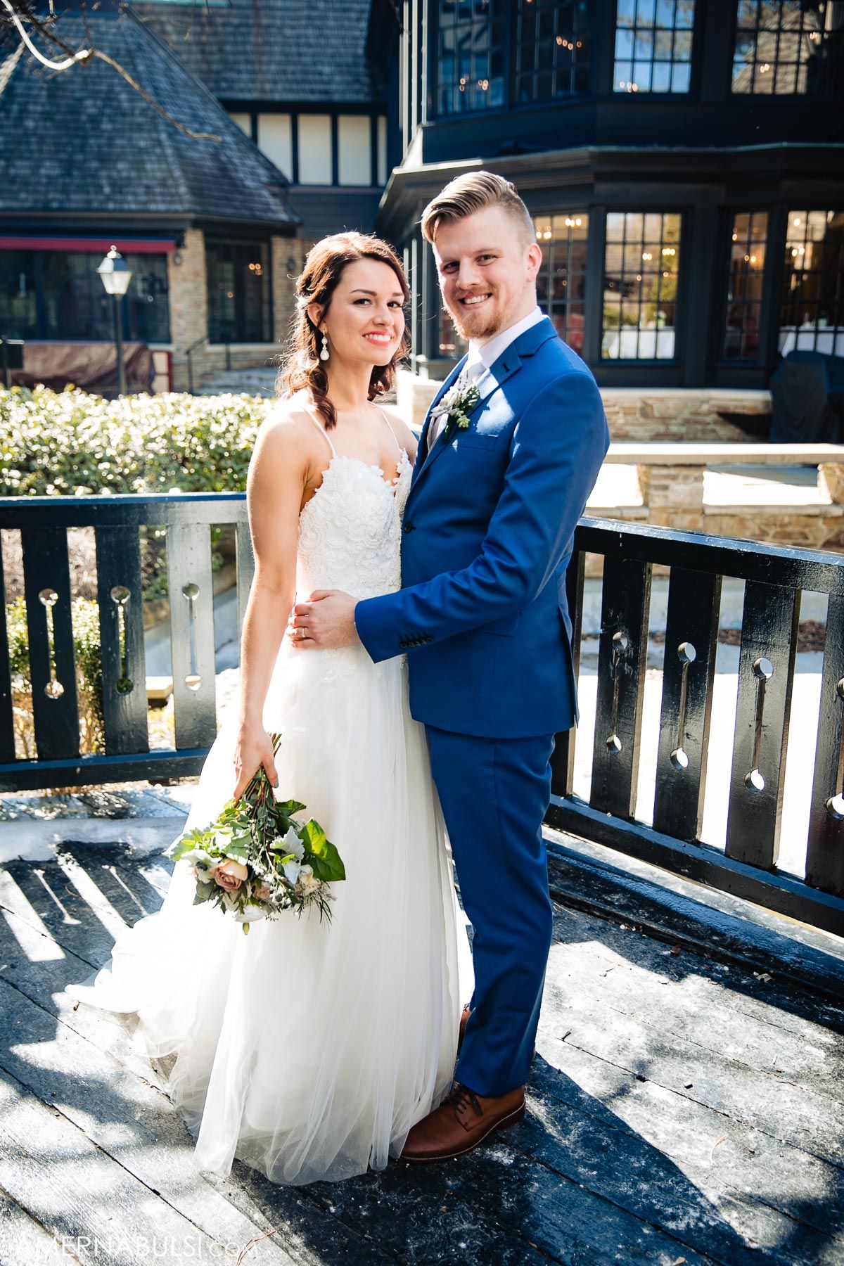 A couple poses outdoors in wedding attire, with the bride in white and groom in a royal blue suit on a sunny day.