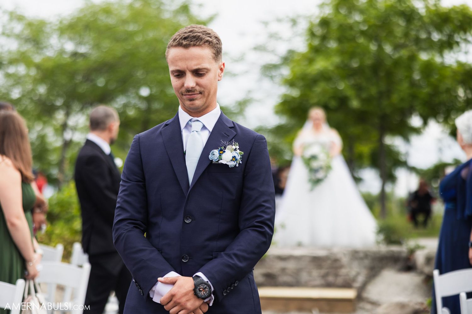 Wedding party poses outdoors with guests in navy and gray attire near green trees.