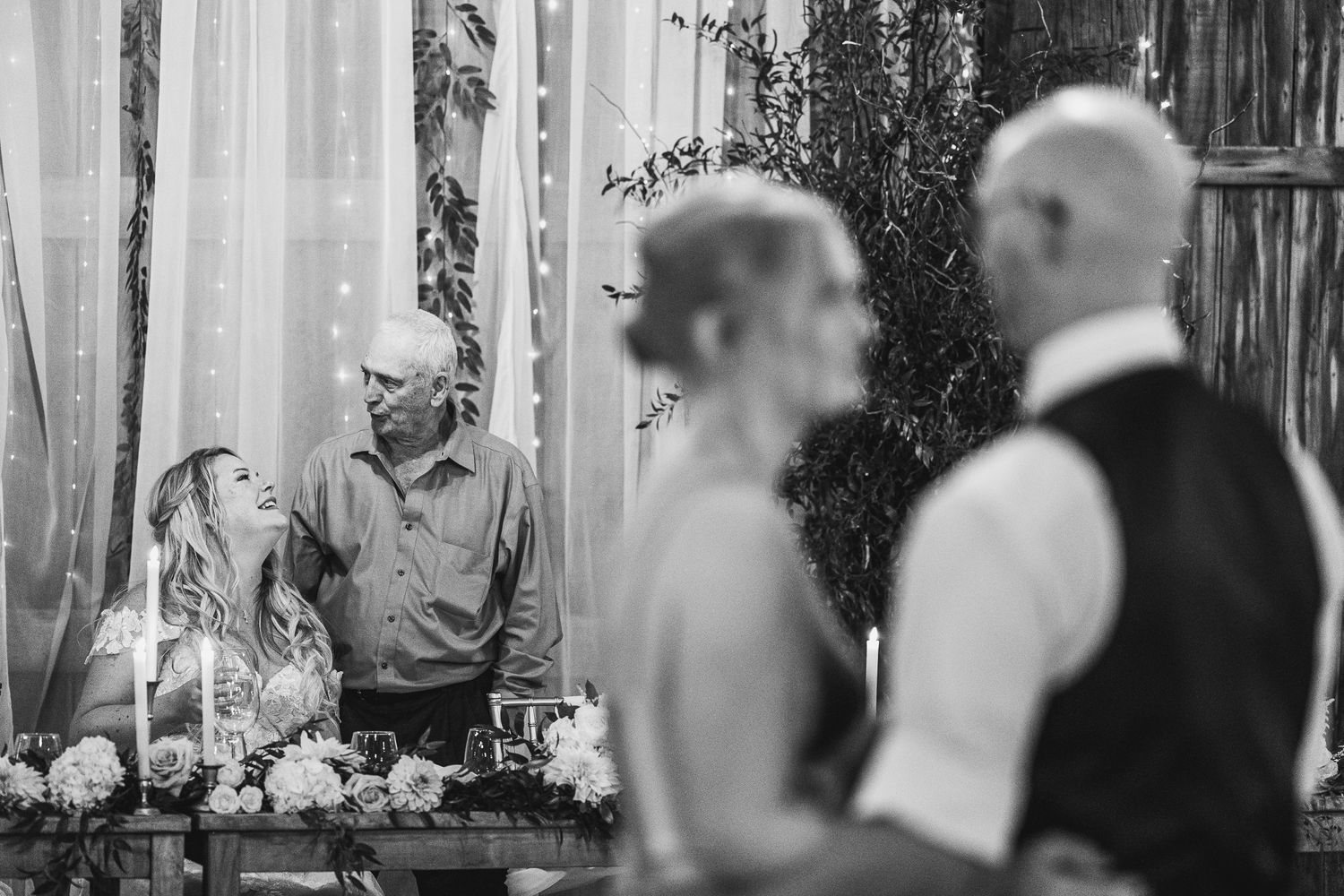 Black and white wedding photo of couples dancing together during a reception in a rustic venue.