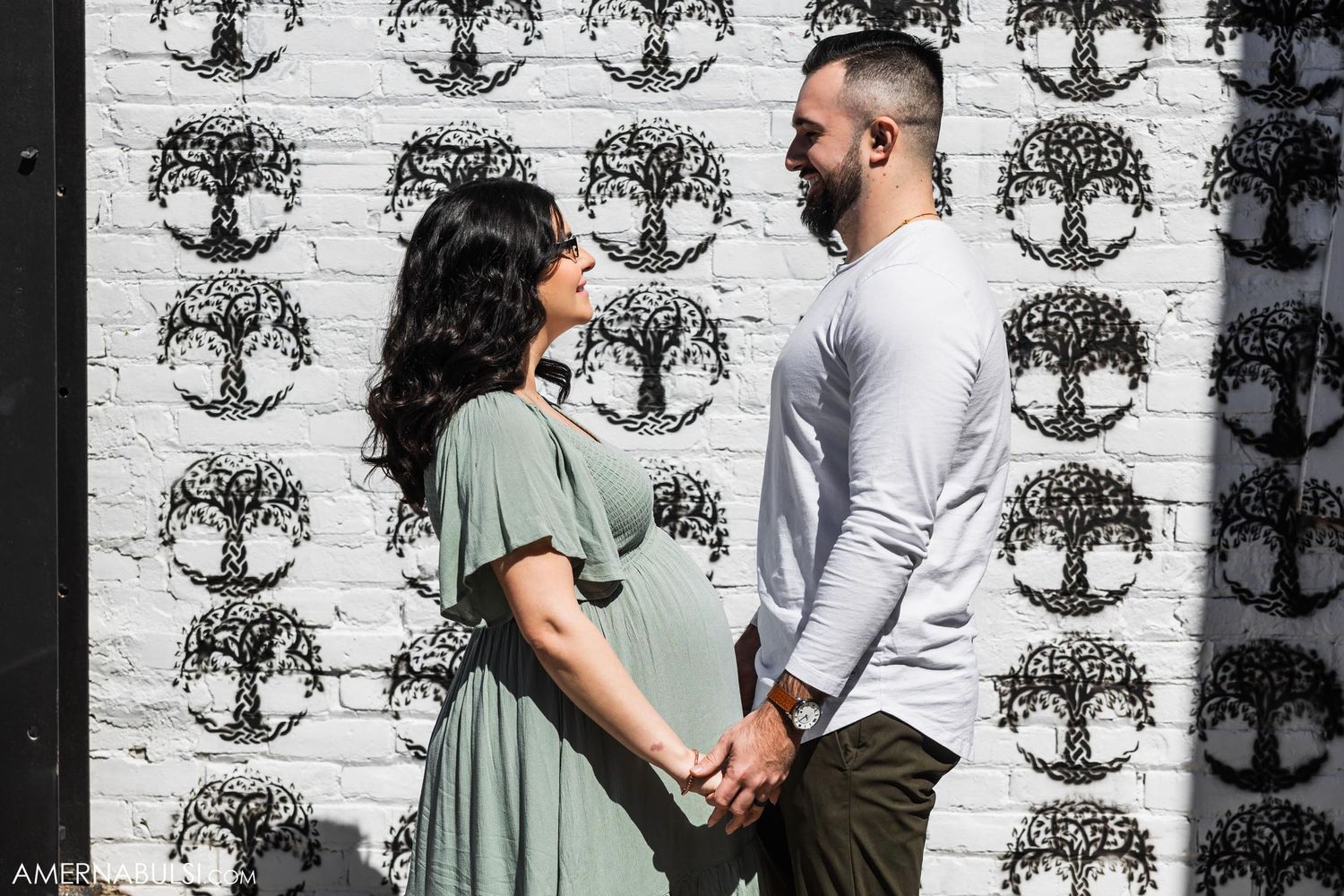 An expectant couple poses against a wall decorated with tree patterns.