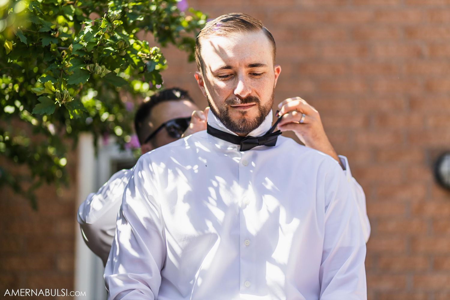 A person in white adjusting their bowtie outdoors near greenery.