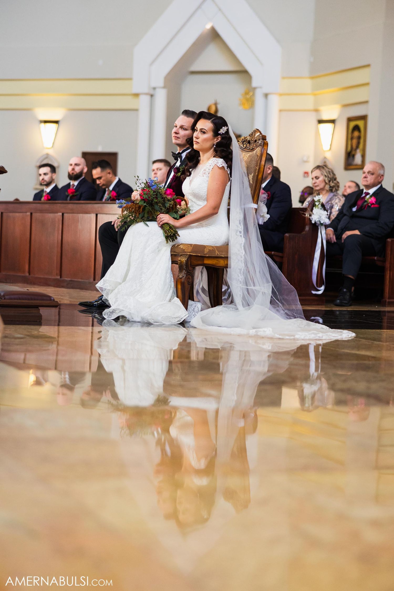 Wedding ceremony in a church with couple seated in front pews with reflective floor and elegant decor.