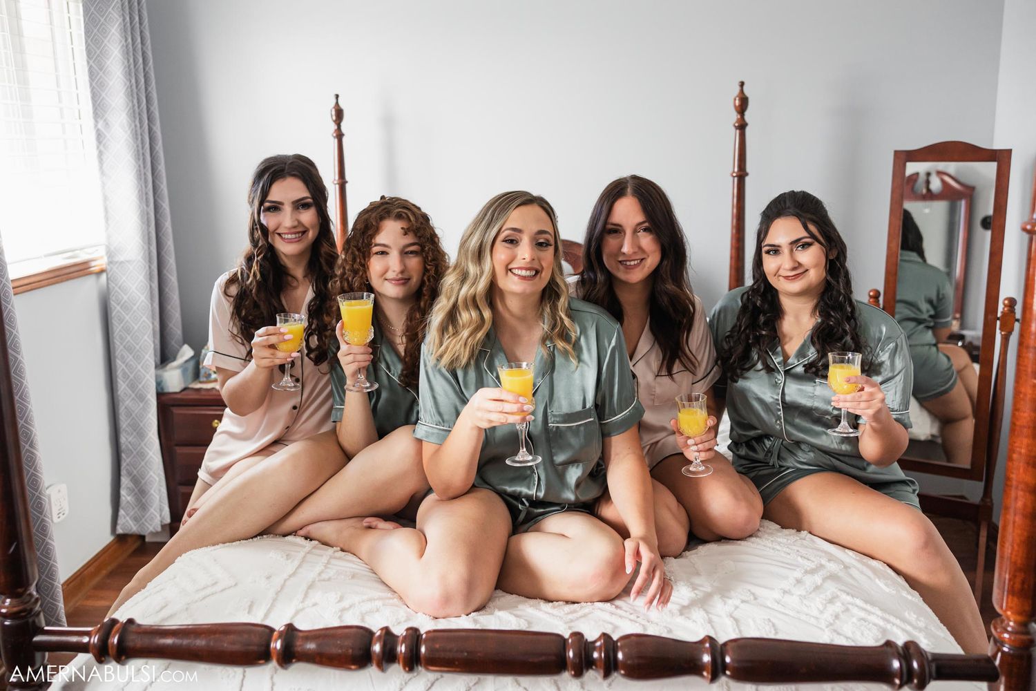 Group wearing matching robes sits on bed while holding mimosa glasses for pre-wedding celebration.