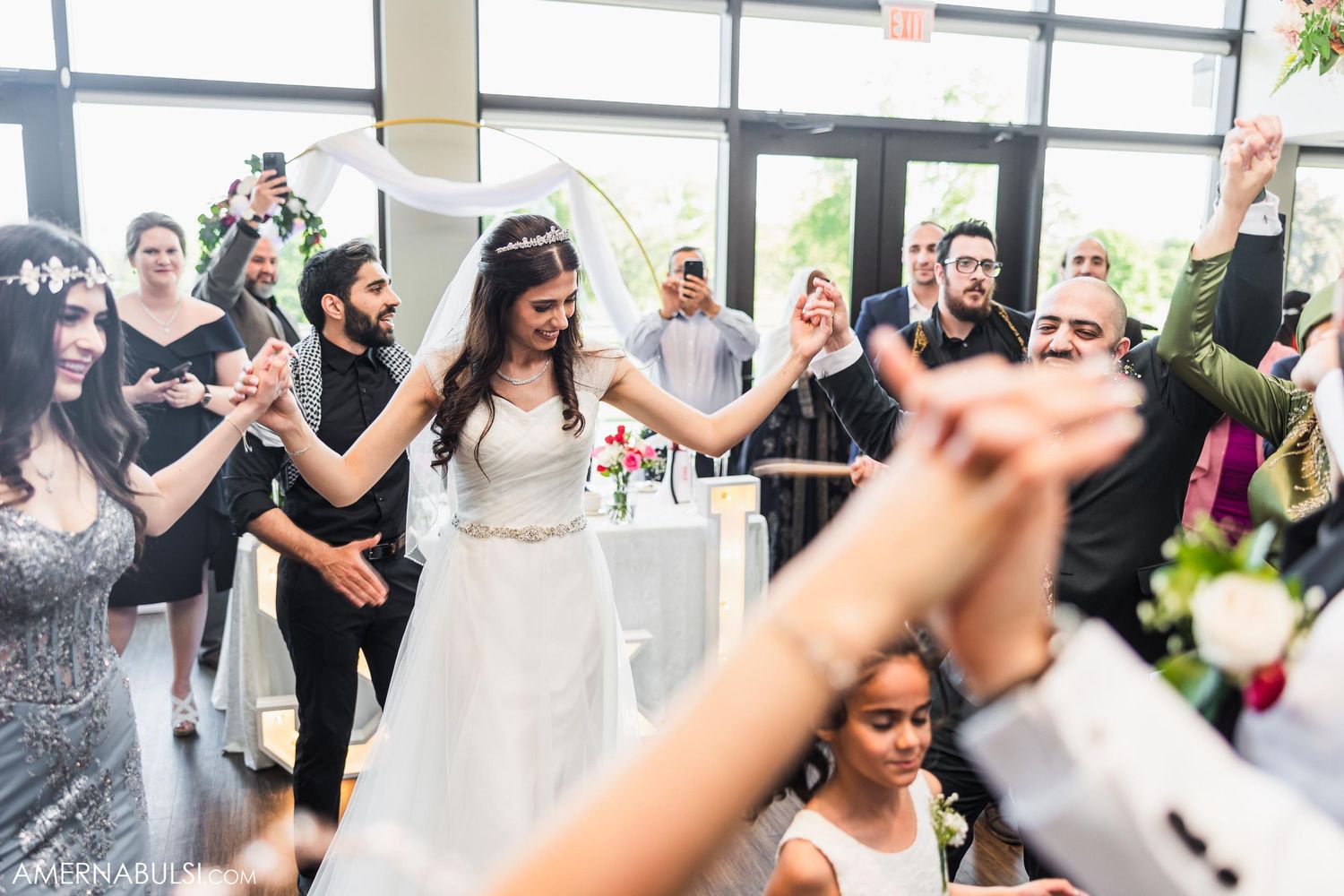 Wedding guests dance and celebrate with raised arms in a bright reception venue.