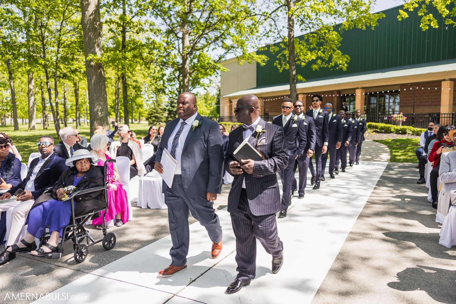 Wedding party in formal attire walking in procession along outdoor pathway.