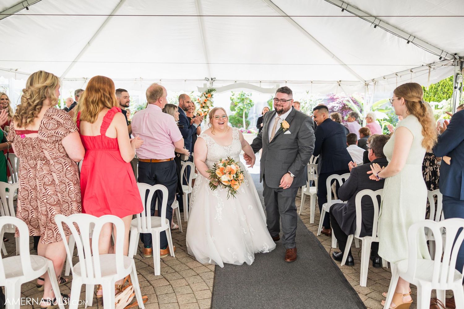 Wedding couple walking down the aisle under a white tent as guests look on during an outdoor ceremony.