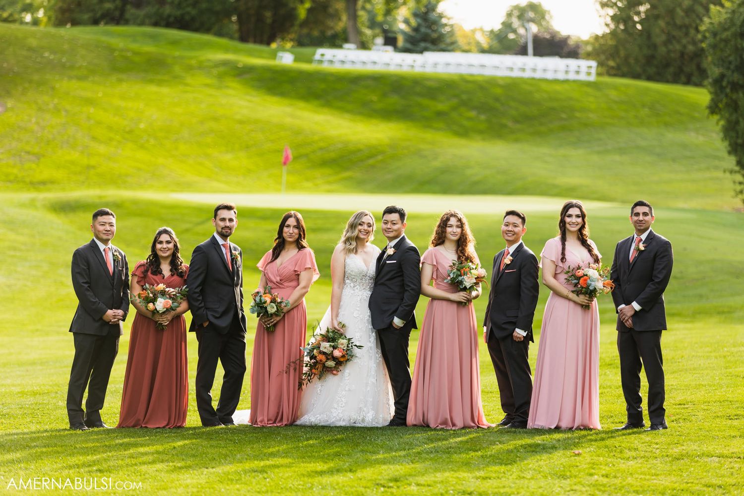 Wedding party in pink dresses and black suits poses on sunny golf course slope.
