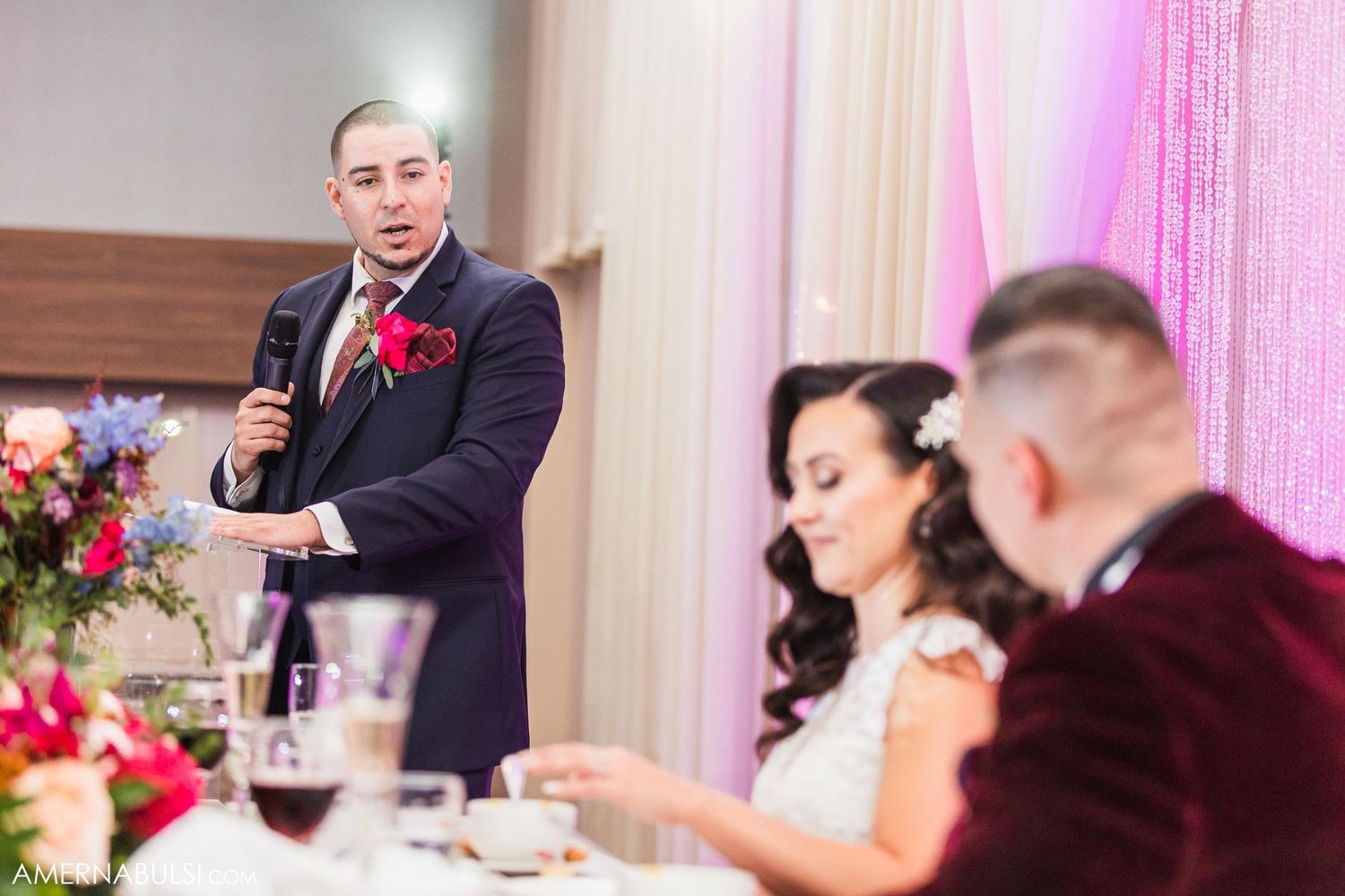 A wedding toast speech being delivered during an elegant reception with pink lighting and floral decor.