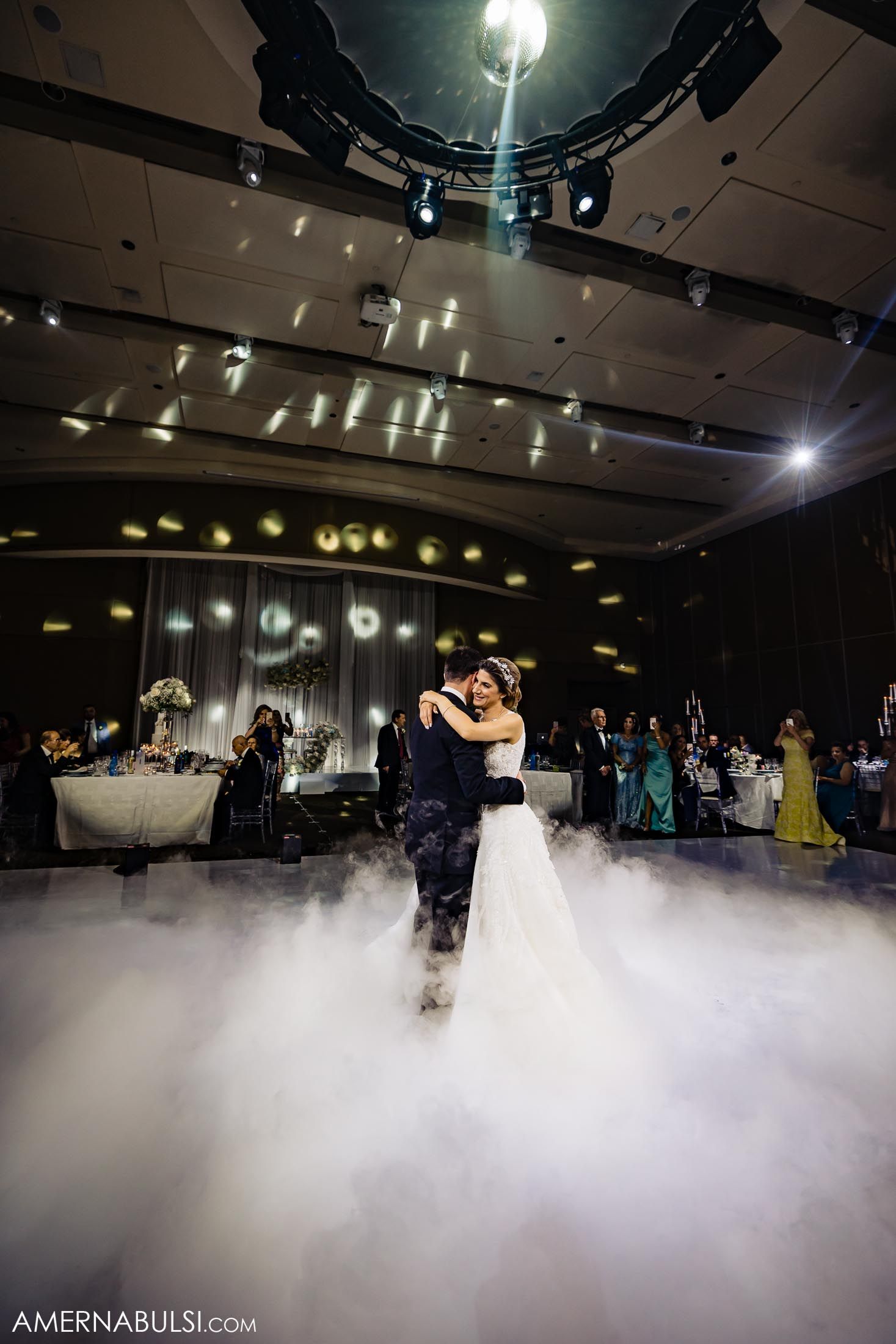 Couple shares their first dance on a fog-covered dance floor under dramatic ballroom lighting.