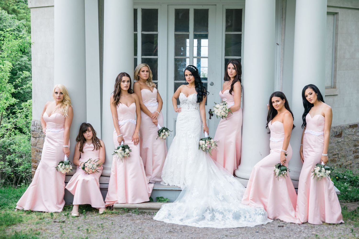 Bridesmaids in pink dresses pose with bride in white gown on mansion steps.